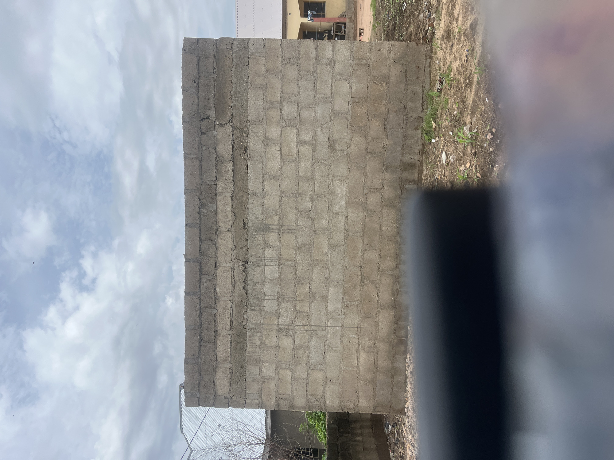 The image shows an unfinished cinder block wall in Jalingo, Nigeria. The wall is the primary subject, with its rough texture and visible mortar lines. To the left of the wall, part of a corrugated metal roof and some greenery are visible. To the right, a yellow building with a white roof can be seen, with a window and clothes hanging outside. The ground in front of the wall is dirt with scattered small stones and some sparse vegetation. The sky above is cloudy. The bottom of the image is obscured by a dark, out-of-focus foreground element, possibly a vehicle or a person's head.