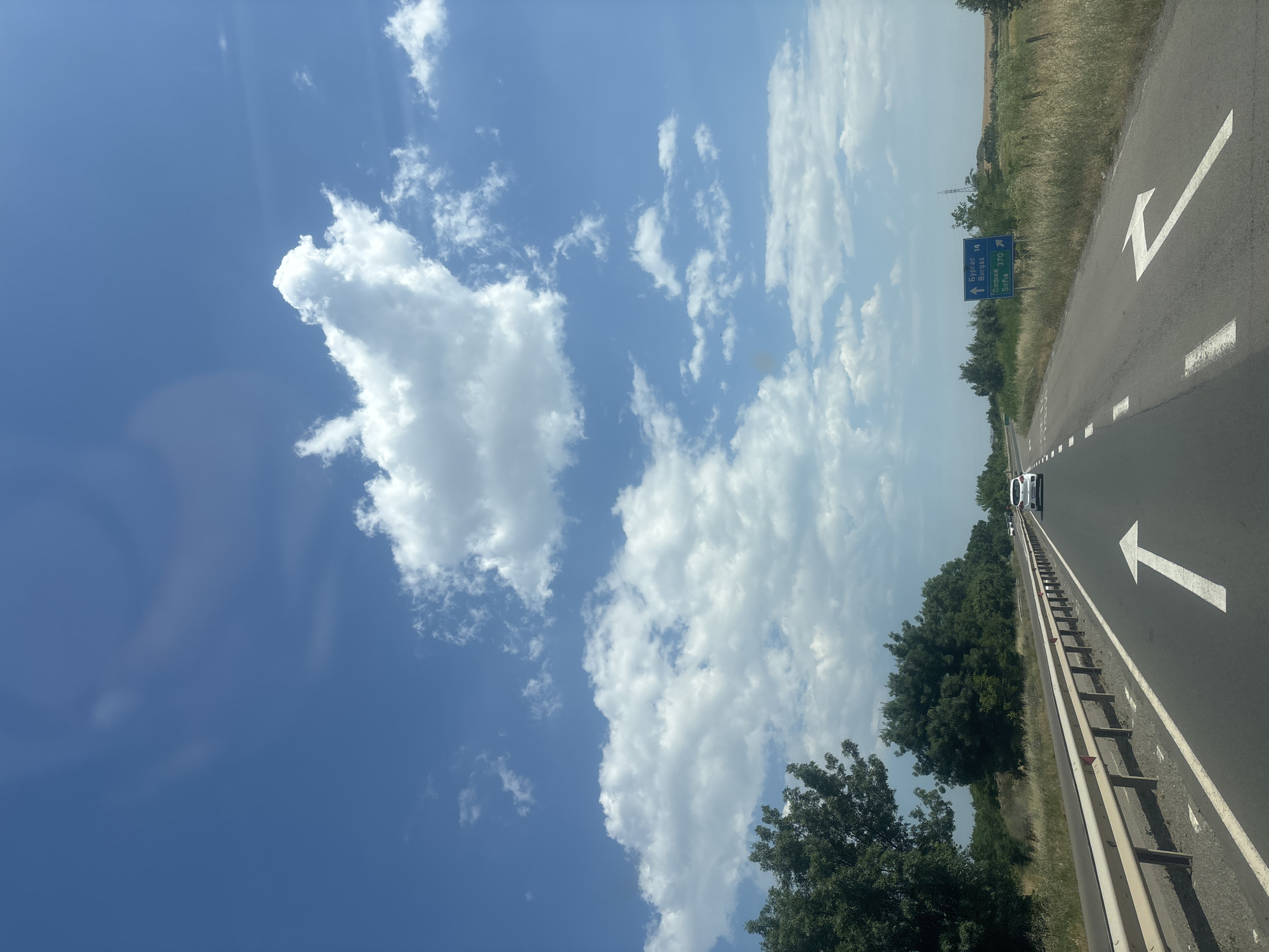 The image shows a view from a moving vehicle on a highway in Burgas, Bulgaria. The sky is a vibrant blue with scattered white cumulus clouds, indicating a sunny day with fair weather. The highway has clear lane markings, including white arrows indicating the direction of travel. A white barrier runs along the side of the road, separating it from the grassy verge and a line of trees. In the distance, other vehicles can be seen on the highway, and a few power lines are visible in the background.
A blue road sign provides directional information. The text on the sign is in Bulgarian and English, indicating distances to "Burgas 14" and "Yambol 370". This suggests the vehicle is traveling away from Burgas and towards Yambol. The presence of trees and dry grass along the roadside hints at a rural or semi-rural setting, possibly during a warm season given the clear sky and vegetation. The overall impression is one of travel on a bright, clear day.
