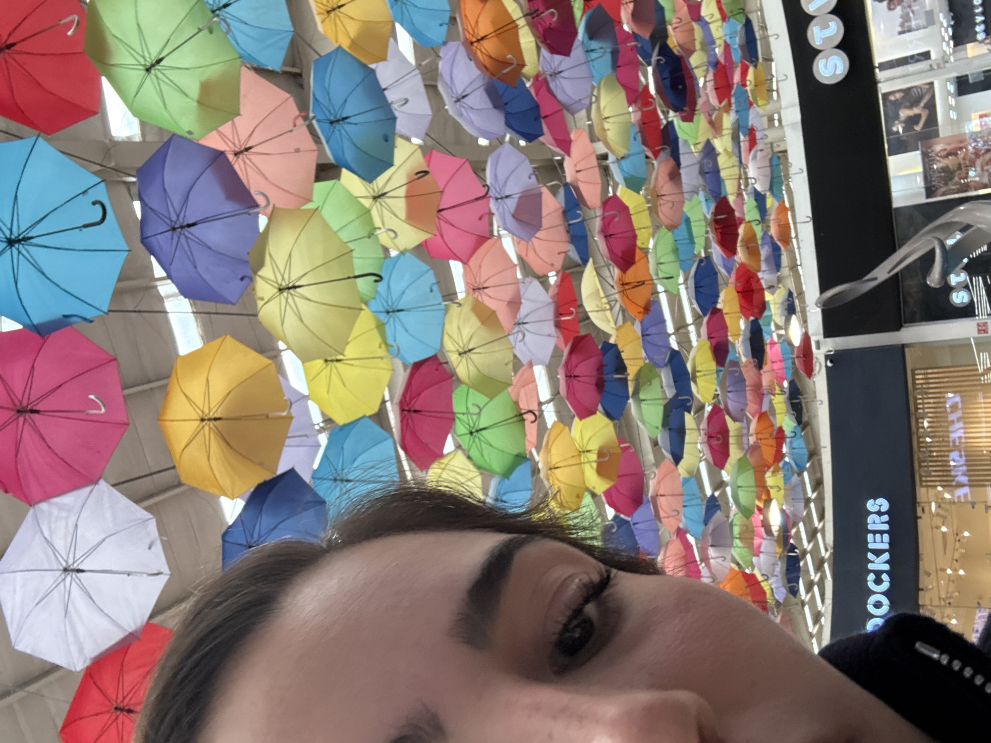 A medium close-up shot shows a person's face in the lower portion of the frame, gazing slightly upward. Above, a dense installation of colorful umbrellas fills the majority of the background. The umbrella colors include red, pink, purple, blue, green, yellow, orange, and white. In the upper-right corner, the vertical text "DOCKERS" is partially visible against a darker surface, suggesting a retail store setting. Other shop signs are partially visible as well. The location appears to be an indoor shopping mall, based on the ceiling structure. It can be inferred that this is not in San Francisco Ocotlán (Ocotlán), Mexico, as the Dockers chain is not a major retailer in that location.