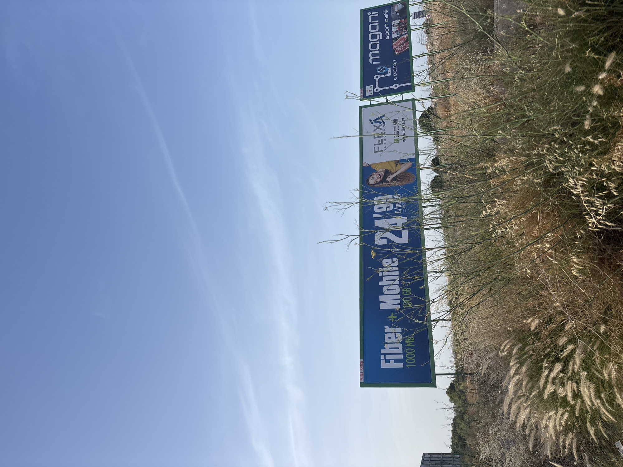 The image depicts a clear blue sky with sparse white clouds over a semi-arid landscape with dry grass and shrubbery in the foreground. In the middle ground, a series of billboards are visible. The left billboard features large blue text reading "Fiber + Mobile" and "24'99" with smaller text indicating "1,000 MB" and "100 GB." To the right of this is a billboard for "FLEXA NETWORKS," displaying a woman's portrait and contact information. Further to the right, a green billboard advertises "magani sport café" with images of food and an address "C/ ENELDO, 3." The overall scene suggests an outdoor setting likely on the outskirts of a city, possibly near a road, given the presence of billboards.