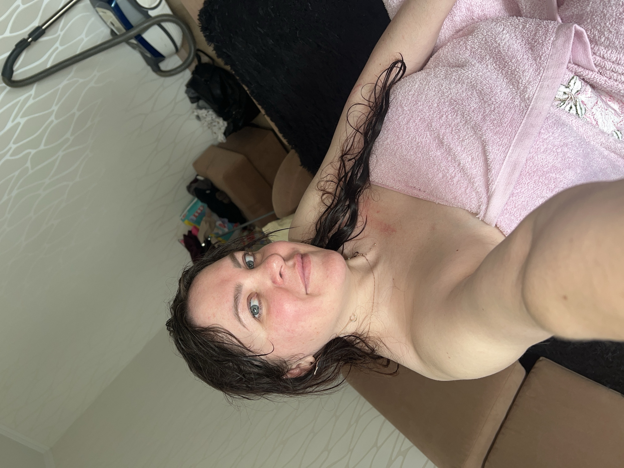 A woman with wet dark hair and fair skin is lying on a brown sofa. She is partially covered with a pink towel. She has light blue eyes and is looking at the camera. She is wearing a gold necklace. The background includes a patterned wall, a black furry object, and a vacuum cleaner. The location is in Perm, Russia.