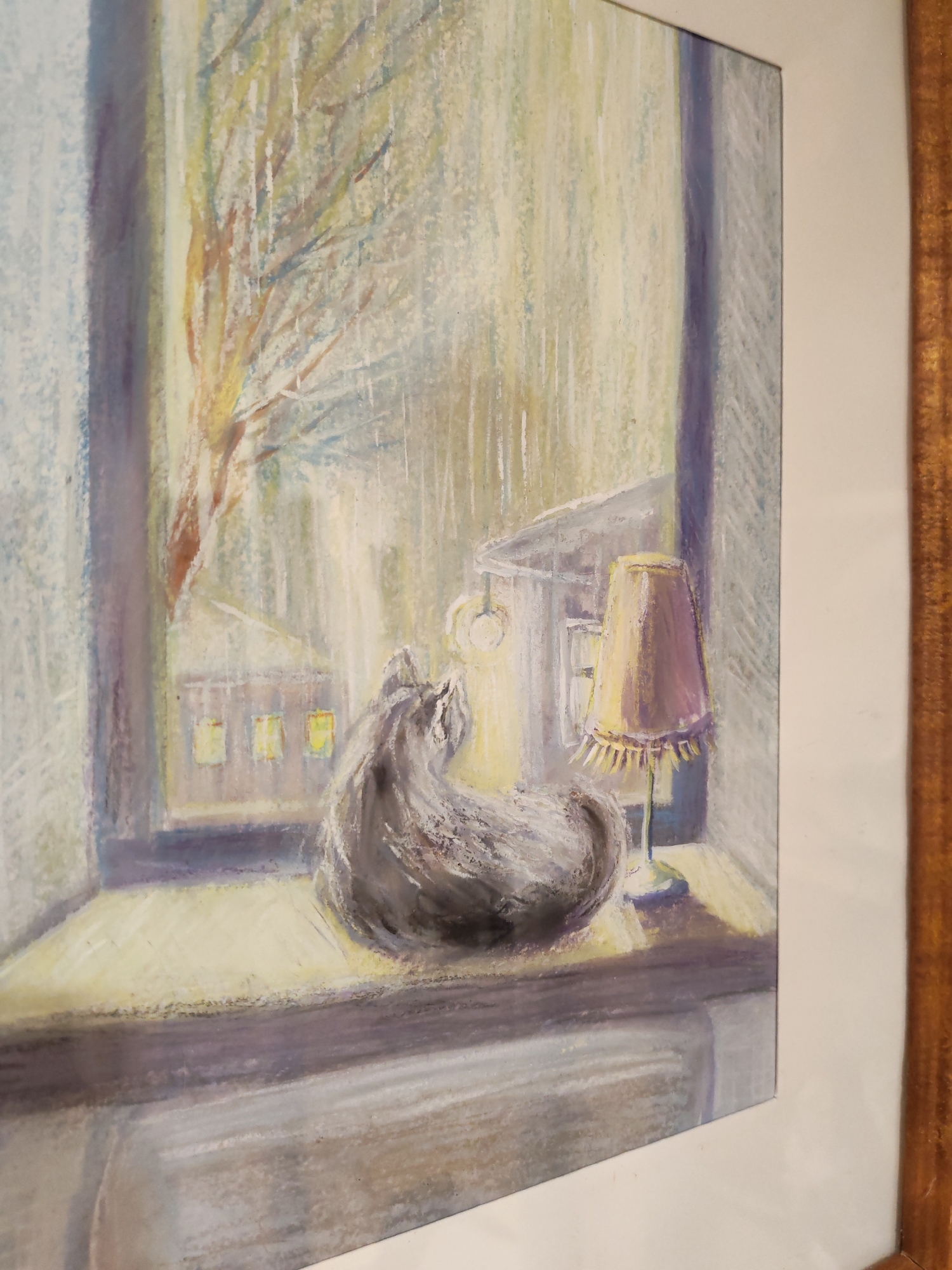 The image shows a framed pastel drawing of a cat sitting on a windowsill looking out at a rainy street scene. The cat is gray and curled up, facing the window. Outside the window, rain is falling, obscuring the view of buildings and a bare tree. A street lamp and a table lamp are also visible in the scene. The drawing is framed with a white matte and a gold-colored wooden frame. There is no indication of the city of Chaikovsky, Russia.