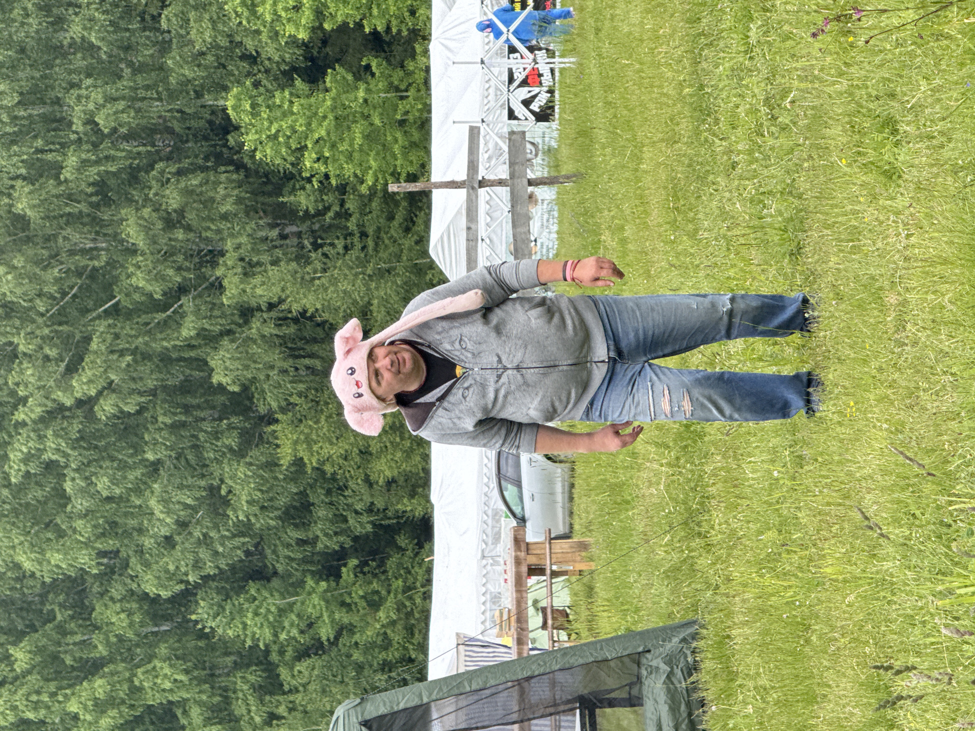 The image, taken in Большая Кивара, Russia, captures an outdoor scene dominated by a man in the foreground. He stands on a grassy field, looking towards the viewer with a slight smile. He is wearing a light grey zip-up hoodie over a dark t-shirt, blue ripped jeans with visible tears on the knees, and a distinctive light pink hat featuring bunny ears, black eyes, and a mouth. Multiple colored bands adorn his right wrist.
In the mid-ground, behind the man, several white canopy tents are set up, indicative of an outdoor event or temporary encampment. A light-colored car is partially visible next to one of these tents. A simple wooden cross-like structure stands between the man and the tents. To the right of the tents, another person wearing a blue jacket is partially visible. The background, particularly on the left, features a dense forest of lush green trees. A portion of a green tent or shelter with a mesh window is visible in the bottom-left corner.
The ground is covered with long, vibrant green grass, interspersed with small yellow and purple wildflowers. The lighting suggests it is daytime, possibly under an overcast sky, creating soft, diffused illumination. Text is faintly visible on one of the white tents in the background, appearing to include letters such as "RK" and possibly "LIVE," though difficult to read clearly. The man's playful hat contributes to a relaxed and possibly festive atmosphere.