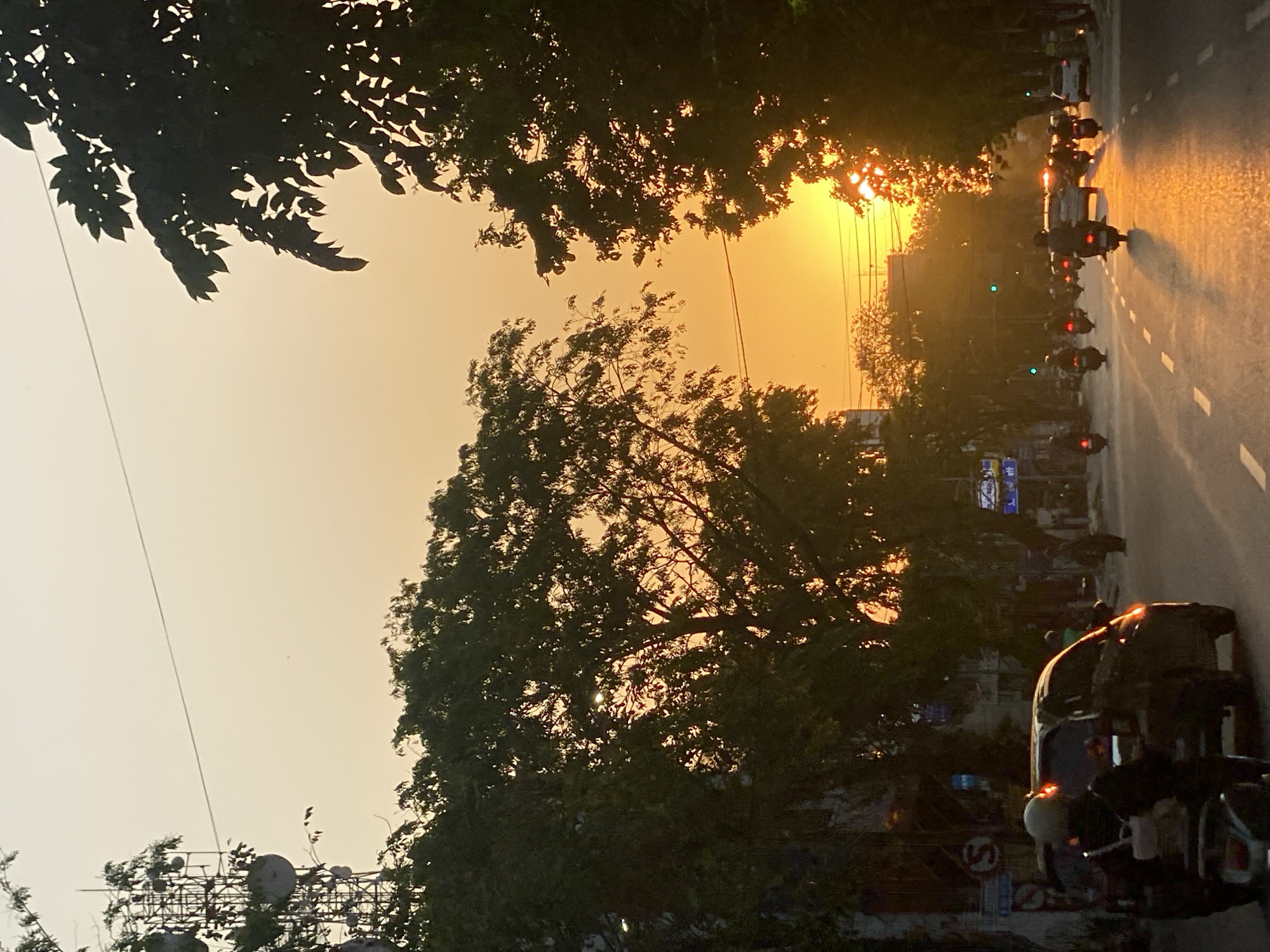This is a photograph taken at sunset on Jalan Lambung Mangkurat in Banjarmasin, Indonesia. The street is paved with asphalt and has lane markings. The sky is a vibrant gradient of orange and yellow, with the sun setting behind silhouetted trees and buildings. Traffic is moderate, with several motorcycles and cars on the road. A tall telecommunications tower is visible on the left side of the image. The overall atmosphere is warm and serene, characteristic of a tropical sunset.