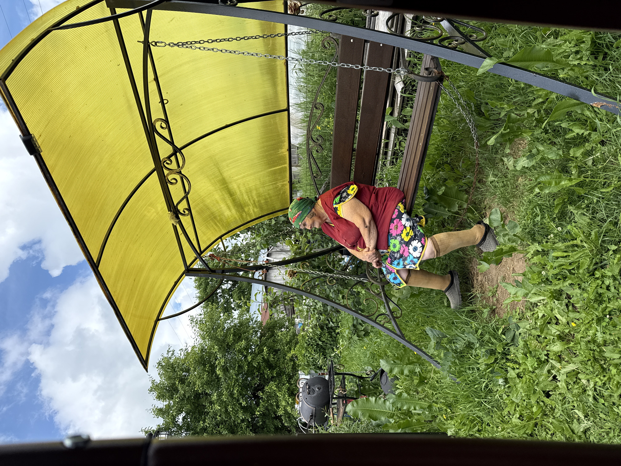 An elderly woman sits on a park swing in a backyard in Zhigalki, Russia. The woman wears a red shirt, a colorful floral skirt, and a green headscarf. She has brown hair and is looking to her left. The swing is under a yellow canopy. The background shows green trees and a wooden fence. A black barbecue grill is visible to the left of the swing. The overall impression is one of a peaceful summer day.