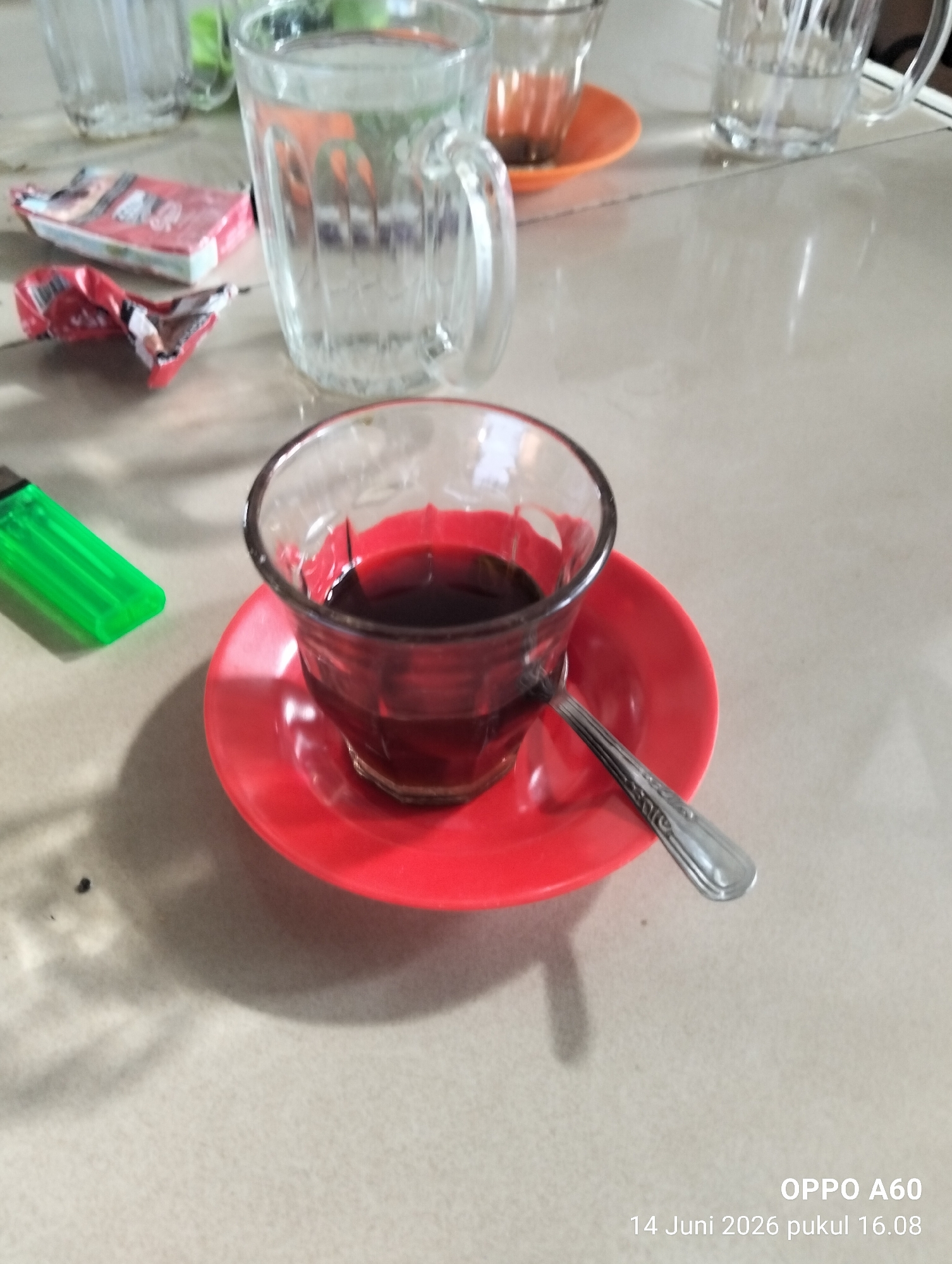The image displays a close-up view of a table setting, likely in Lhokseumawe, Indonesia. The primary focus is a small glass of dark liquid, presumed to be coffee, resting on a bright red saucer. A silver spoon with decorative etching lies on the saucer, partially submerged in the coffee. To the left of the glass, a green plastic lighter is visible. In the background, several other glasses, one filled with a clear liquid, are present, along with a crumpled red cigarette pack. The table surface appears to be a light-colored, textured material. The lighting suggests an indoor environment, possibly a cafe or a home. The timestamp at the bottom right of the image indicates "OPPO A60 14 Juni 2026 pukul 16.08", which translates to "OPPO A60 June 14, 2026 at 4:08 PM". There are no people visible in the image.