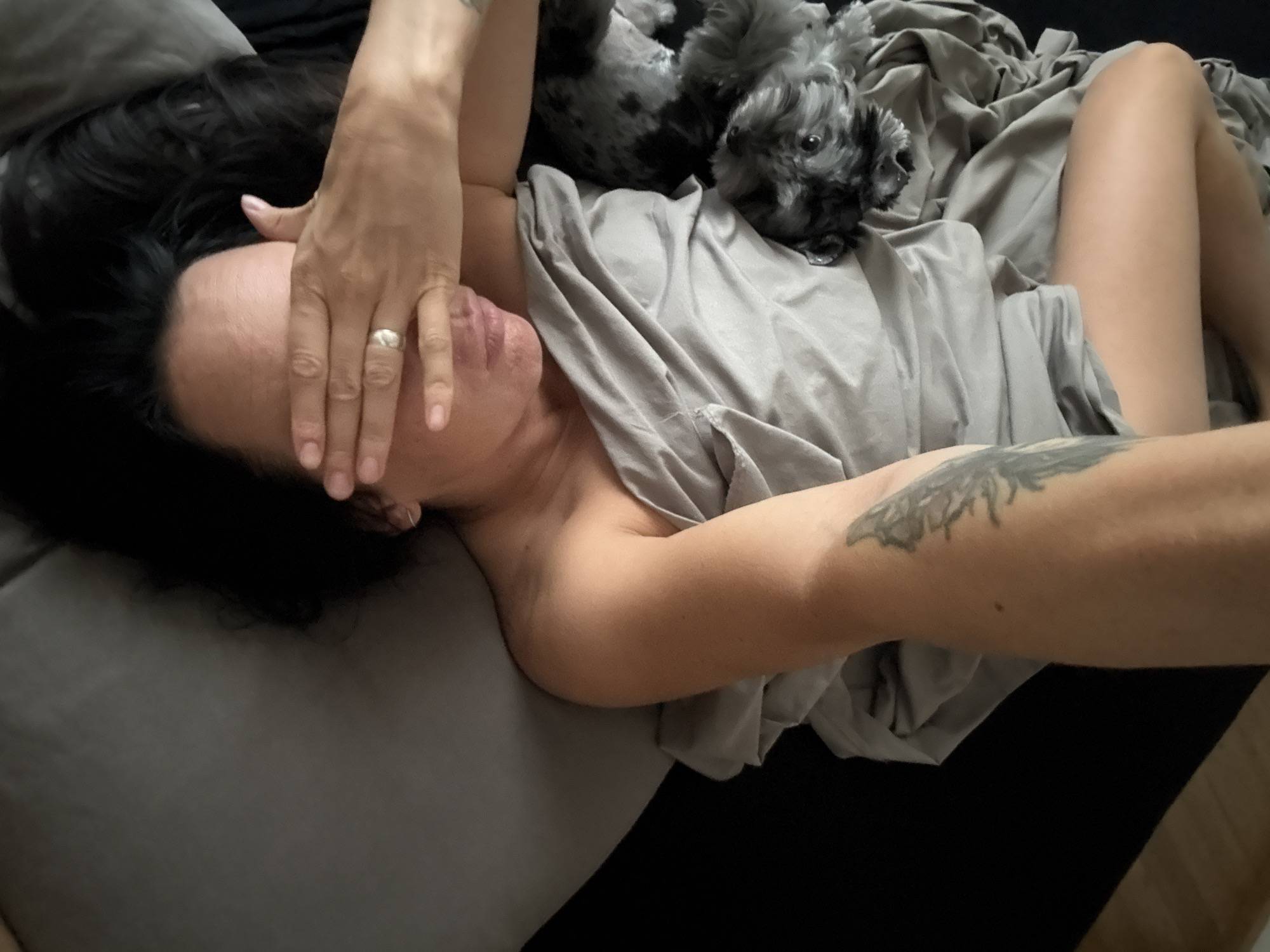 A person is lying on a bed with a small dog. The person is covering their face with their right hand, wearing a ring on their left hand. They have dark hair and tattoos on their arm. The dog is gray and black with curly fur and appears to be lying on the person's chest or stomach, partially covered by a gray blanket. The setting appears to be a bedroom, with dark bedding and a hint of wooden flooring visible. The lighting suggests it could be daytime or a room with artificial lighting. The overall mood is relaxed and intimate.
