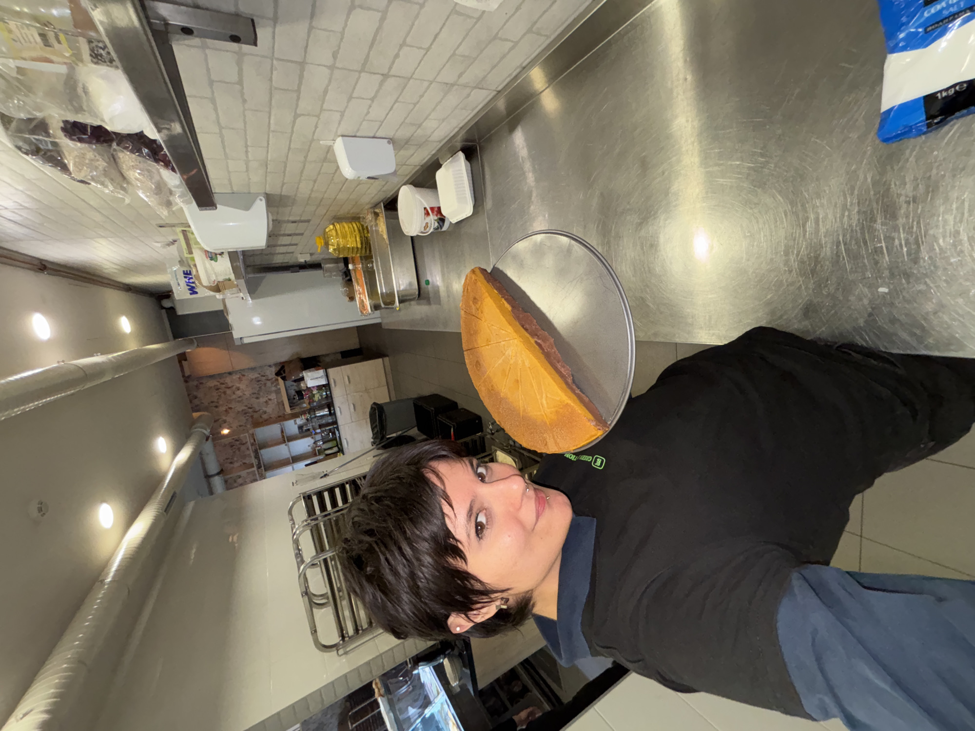 A person wearing a black shirt with blue sleeves is standing in a commercial kitchen, holding a large round piece of food on a silver platter. The kitchen has stainless steel surfaces, white-tiled walls, and fluorescent lighting. There are containers, possibly food storage, on the counter, and a bag of salt is visible in the upper right corner of the image. The person is facing the camera, appearing to pose with the food item. The scene is not identifiable within the city of Sofia, Bulgaria, as there are no landmarks.