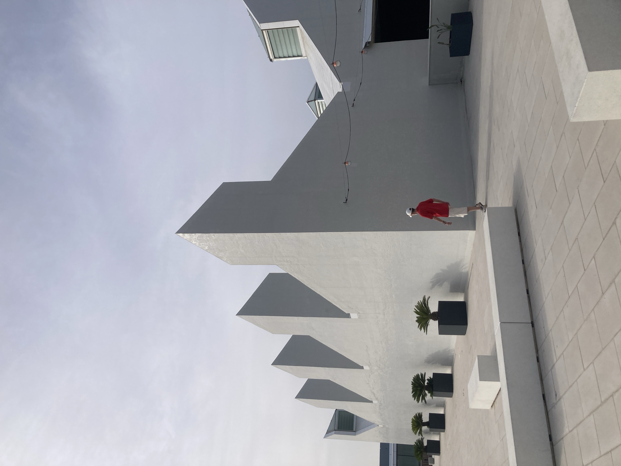 The image shows a person in a red shirt and white shorts walking on a paved plaza in front of a modern building with a jagged roofline. The building is light gray and features geometric shapes. Several potted plants are lined up against the wall, and there are concrete benches on the plaza. String lights are visible along the side of the building. The sky is overcast. The scene is located at the Disseny Hub Barcelona, a multi-purpose building housing several design and fashion institutions.