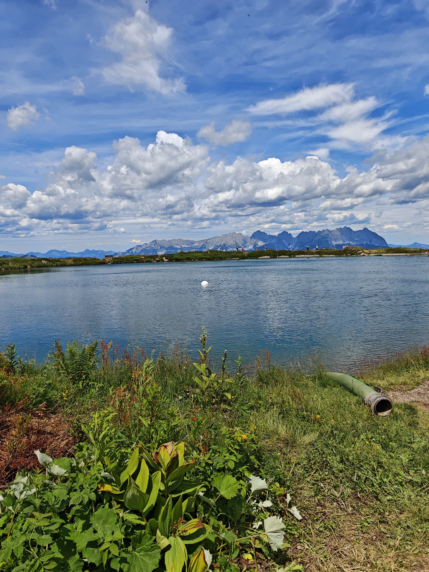 This is a landscape photograph taken on a sunny day with scattered clouds in the sky. The scene depicts a serene lake with mountains in the background. In the foreground, lush green vegetation and wild grasses line the edge of the lake. A large, green pipe emerges from the grass on the right side of the frame, partially submerged in the water.
In the mid-ground, the calm surface of the lake reflects the blue sky and white clouds. A white buoy is visible in the water. Across the lake, the distant mountains rise majestically, their peaks possibly snow-capped. Along the lakeshore in the background, several small figures of people can be seen, some walking or standing, suggesting a leisurely day out. A small building or structure is also visible on the far shore near the mountains.
The overall impression is one of natural beauty and tranquility. The bright sunlight and clear sky indicate pleasant weather, likely mid-morning or afternoon. The location is suggested to be Kirchberg in Tirol, Austria, known for its mountainous terrain and natural lakes. No visible text is present in the image.
