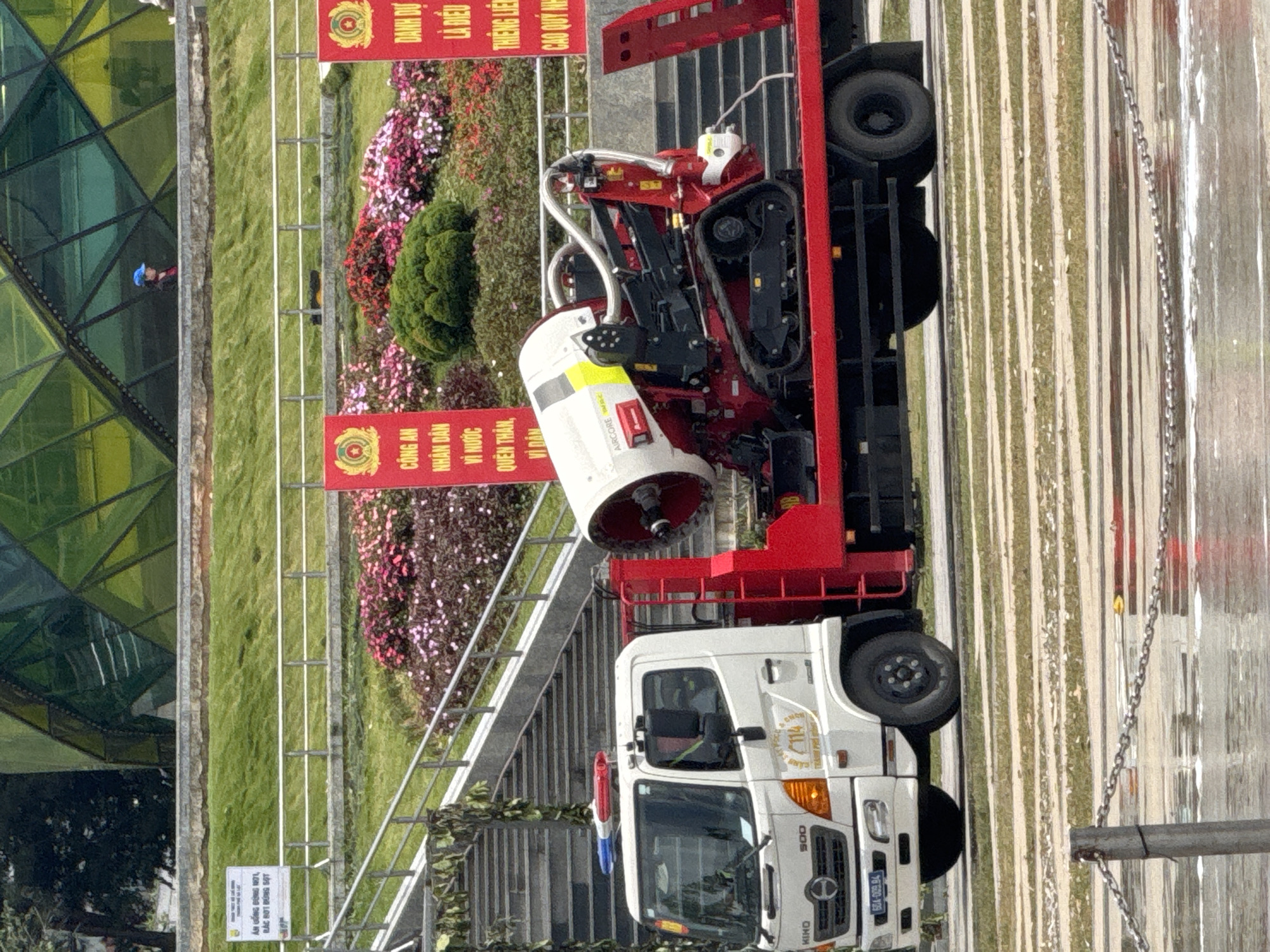 A white Hino truck with a red trailer is parked on a paved surface. The trailer carries a large, red and white fog cannon machine with caterpillar tracks. The truck has blue and red flashing lights on its roof. Two red banners with gold emblems are visible in the background, one on the left and one on the right. The banner on the left reads "AN CÔNG AN NHÂN DÂN VI NƯỚC QUÊN THÂN, VI DÂN" and the banner on the right reads "DANH DỰ LÀ ĐIỀU THIÊNG LIÊNG CAO QUÝ NHẤT". A person wearing a blue cap and a red jacket is standing on a grassy slope behind a stone wall. In the far background, a large, modern building with a green, angular glass facade is visible. The scene appears to be in an outdoor public space, possibly a park or event area, in Da Lat, Vietnam. The overall atmosphere is clean and orderly.