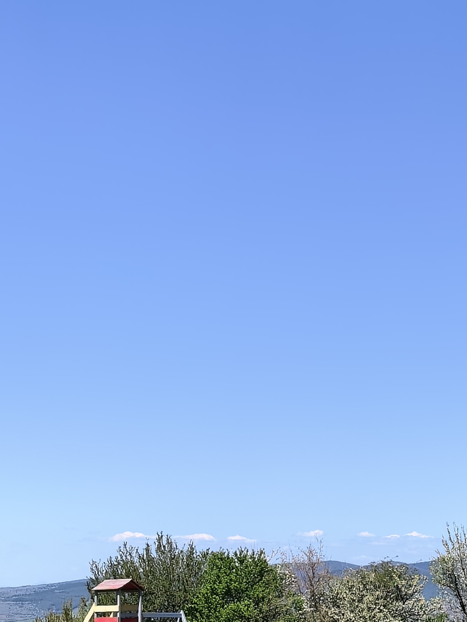 The image shows a clear blue sky above a landscape. In the lower part of the frame, there is a row of green trees and bushes, with a playground structure visible behind them. The background includes a hazy mountain range and what appears to be a cityscape or a suburban area. The city and exact location remain unknown.
