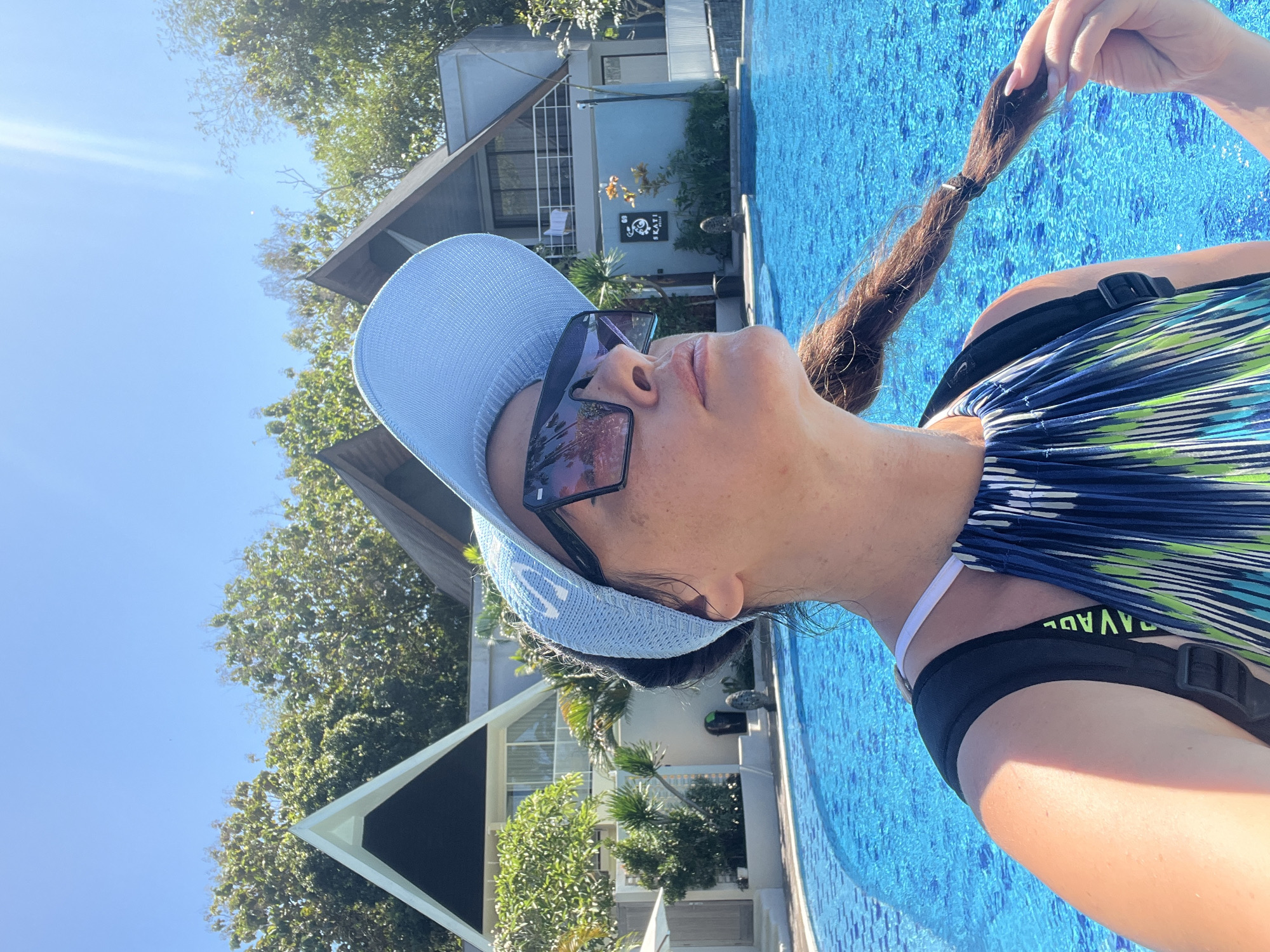 A woman with a braid of dark hair is posing for a selfie by a bright blue swimming pool. She is wearing a light blue baseball cap, sunglasses, and a colorful striped halter top. Behind her, two modern buildings with triangular roofs and a lush green landscape are visible under a clear blue sky with sun glare. The scene appears to be at a resort or hotel in Kuta Selatan, Indonesia.