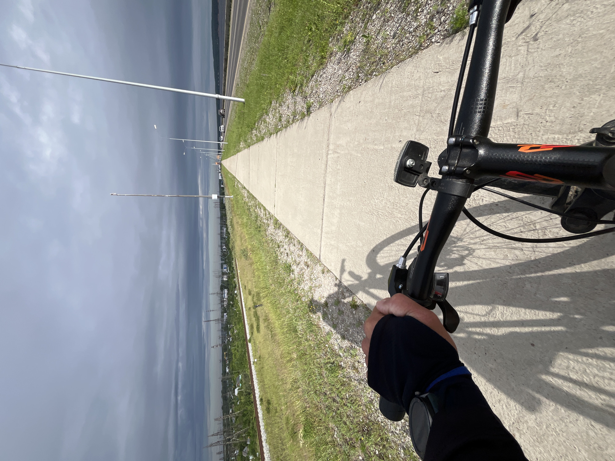 A first-person perspective shot captures a cyclist riding on a paved path alongside a road on an overcast day. The cyclist's gloved left hand is visible on the handlebars of a black bicycle, which is equipped with a front light. The path runs parallel to a grassy area and a set of railway tracks on the left, with a highway featuring several cars in motion on the right. In the distance, a cityscape with numerous industrial towers and buildings is discernible under a heavy, gray sky, likely within or near the city of Chaikovsky, Russia, given the landscape. The overall scene suggests an outdoor recreational activity near an industrial area.