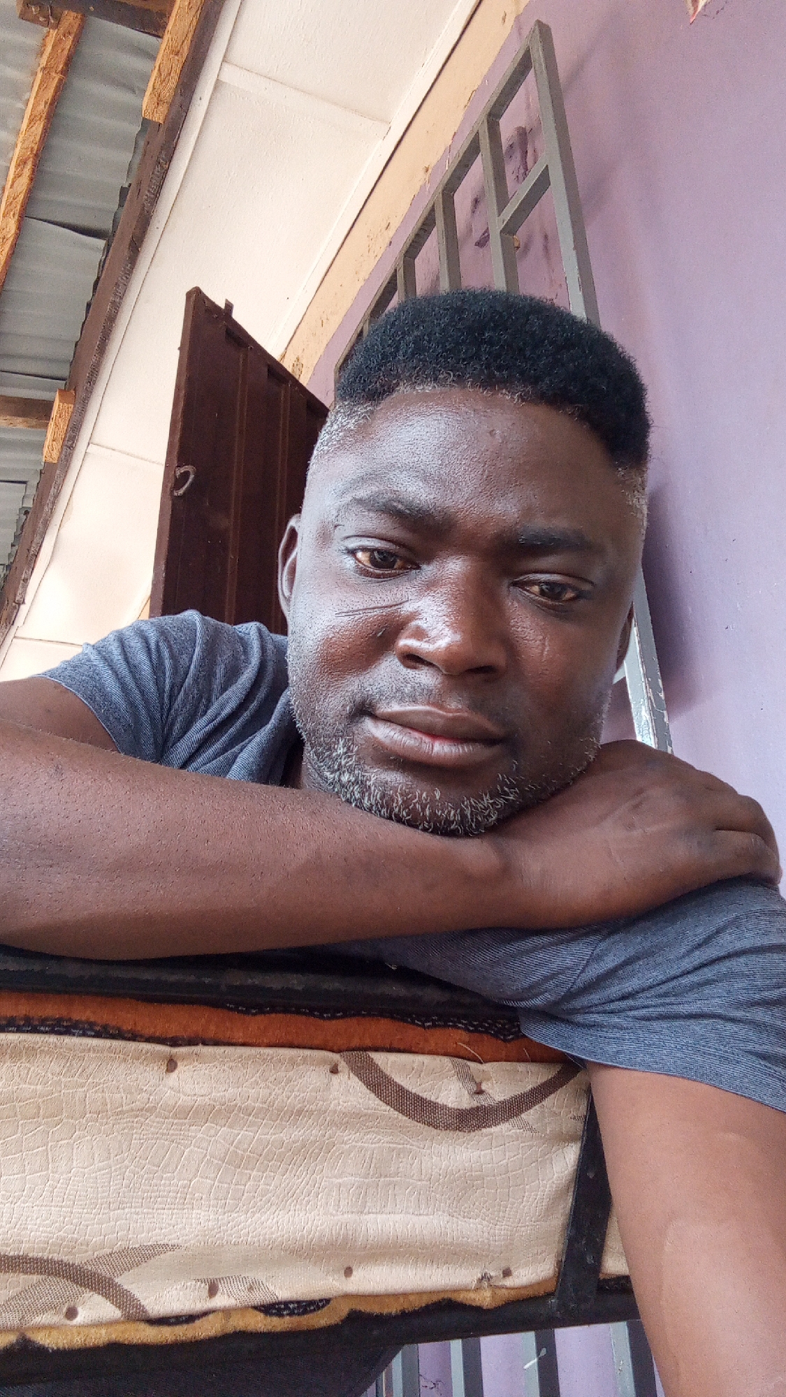 A close-up shot features a man resting his arms on what appears to be a worn-out piece of furniture or a mattress, possibly a chair seat. The man has dark skin, close-cropped hair, and a salt-and-pepper beard. He is wearing a gray t-shirt. Behind him, there is a wall painted in a light purple color, a window with metal bars, and part of a wooden structure that may be a doorway or a roof overhang. Based on the prompt, this is located in Sabon Tasha, Nigeria.