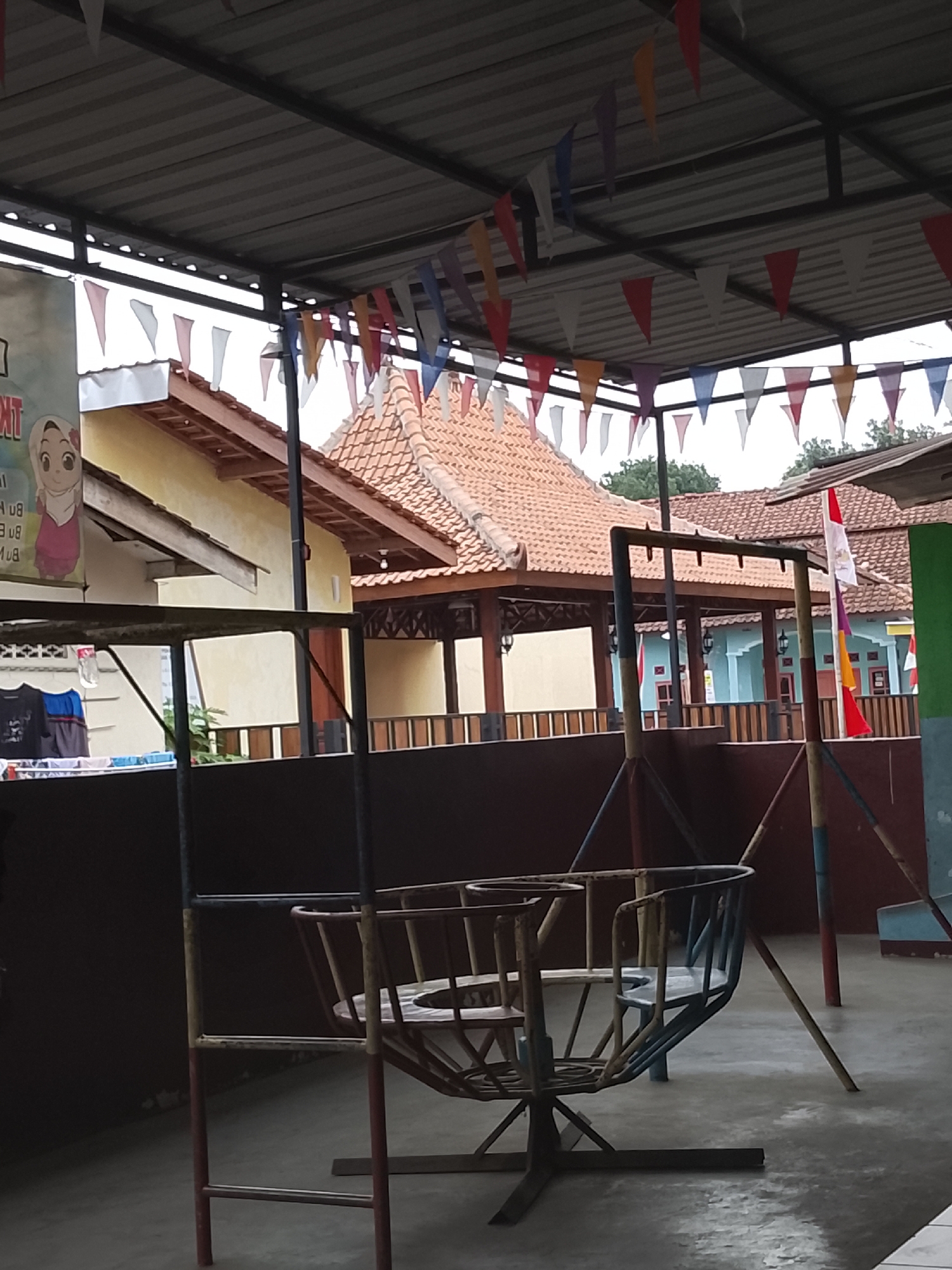 The image depicts an outdoor recreational area under a covered roof supported by metal beams, likely at a kindergarten or community center in Karangkemiri, Indonesia. Colorful triangular flags are strung across the roof. In the foreground, a metal swing set with a circular seat is visible. Behind the swing set, there's a low wall, and further back are buildings with tiled roofs. On the left, a banner featuring a cartoon character wearing a hijab is displayed. Indonesian flags are also visible hanging on poles. The ground is paved with concrete. There are no people visible in the image.