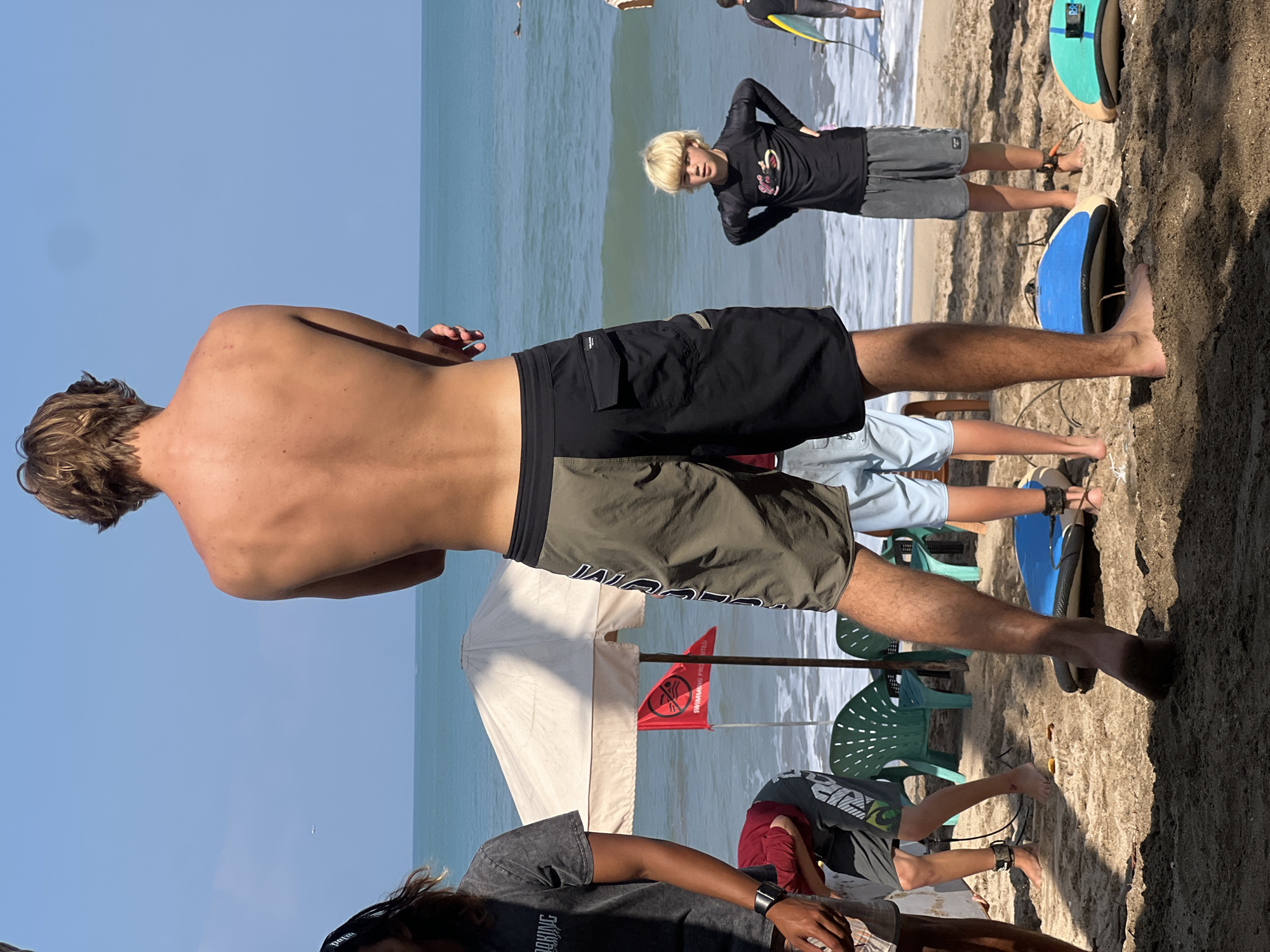 The image depicts a sunny beach scene on a clear day, likely in Kuta, Indonesia, given the context. The primary focus is on a tall, fair-haired young man facing away from the camera, wearing two-toned board shorts. He stands on a sandy beach near the ocean, where small waves are breaking. Several other individuals are present, including a younger boy with blonde hair in a black rash guard and grey shorts, standing closer to the water. Other people are partially visible, some wearing surf gear. There is a white beach umbrella with a red "swimming prohibited" flag, suggesting potentially hazardous conditions. Several surfboards are lying on the sand. The overall atmosphere is that of a surf-friendly beach, possibly a surf school or a popular surf spot.