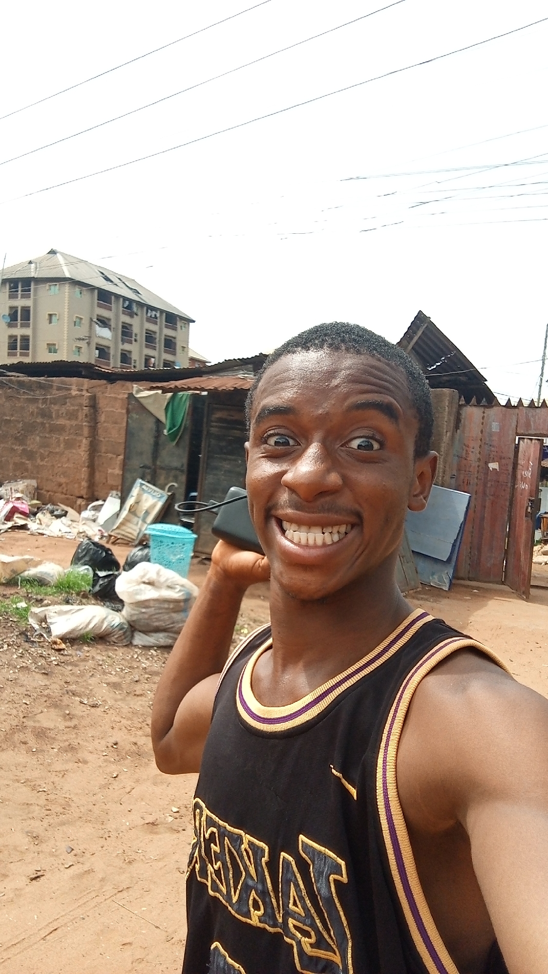 A young man is taking a selfie outdoors in Onitsha, Nigeria. He wears a black Lakers tank top with yellow trim. He has short black hair and is smiling broadly, holding a black object to his ear as if on a phone call. Behind him, there is a collection of trash bags and discarded items on a dirt ground. A brick wall and corrugated metal structures are also visible. In the background, a multi-story apartment building is present, along with overhead power lines. The environment appears somewhat impoverished and unkempt.