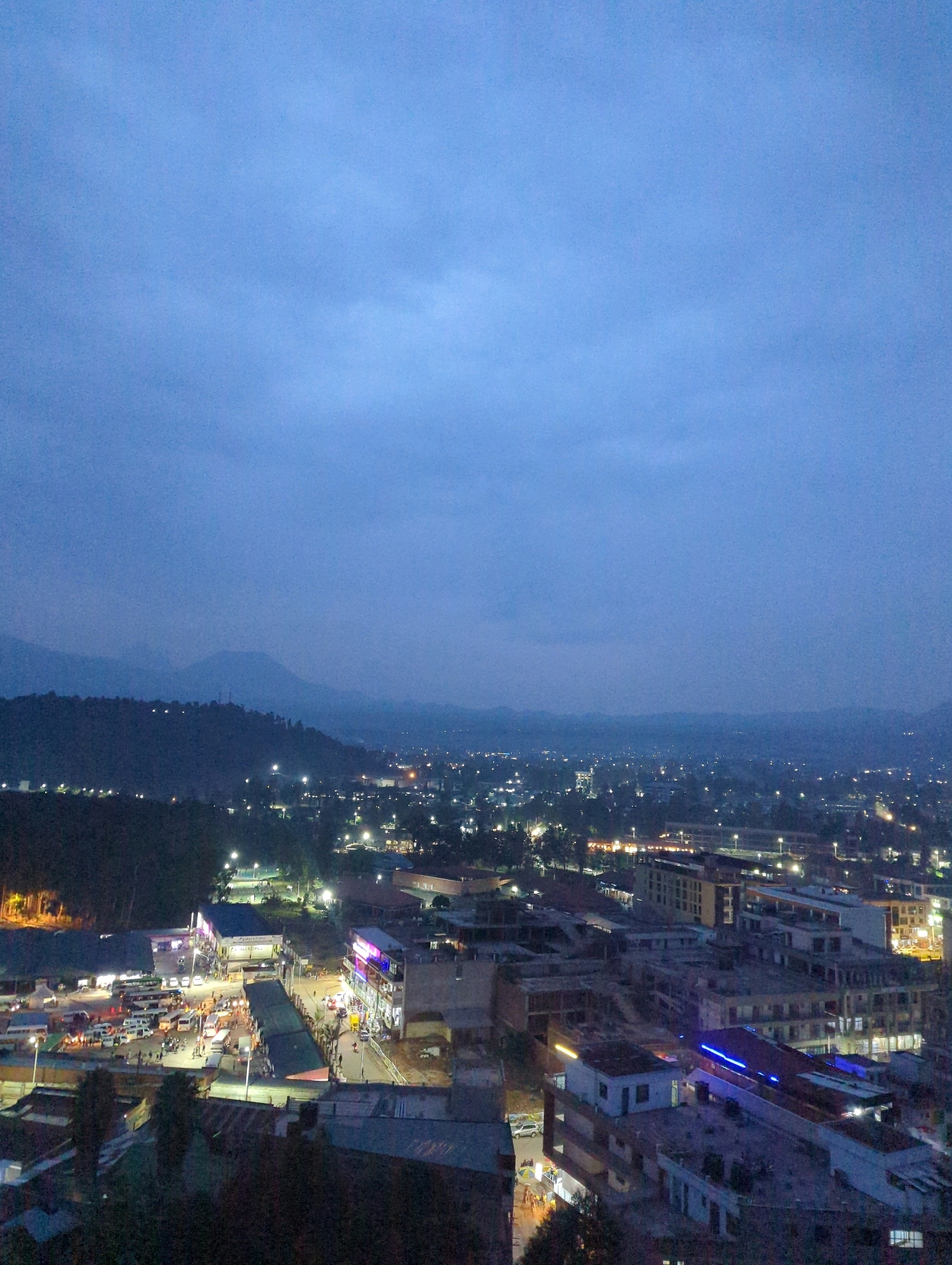 The image is a high-angle, long-shot taken at dusk in Musanze District, Rwanda. The sky is a deep, muted blue, indicating twilight. In the foreground, a dense cluster of buildings with lit windows and streetlights illuminates the urban landscape. Some buildings appear to be under construction. A busy street or market area is visible, with numerous vehicles, including vans and buses, parked or moving. Pedestrians are present in small groups around the market stalls and buildings.
The midground features a hillside covered in trees and dotted with scattered lights, suggesting more residential areas or smaller settlements on the outskirts. Further in the distance, rolling hills and mountains are silhouetted against the fading light, with a hazy, atmospheric effect obscuring finer details. The overall impression is of a developing city at the transition from day to night, nestled within a mountainous region. The lighting suggests a cool, slightly humid environment typical of this region.