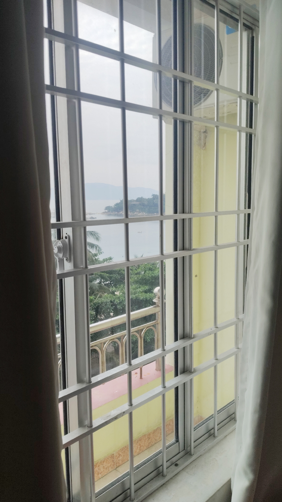A view from a window with white bars reveals a coastal scene. The window frame is also white. An air conditioning unit is visible outside the right side of the window. Beyond the window bars, a body of water extends towards a landmass. Trees and greenery are also visible below and to the left, along with a balcony with decorative railings. A pink surface can be seen through the window. A pale curtain is draped to the left of the window, and another curtain to the right. The location is Bac Nha Trang Ward, Vietnam.