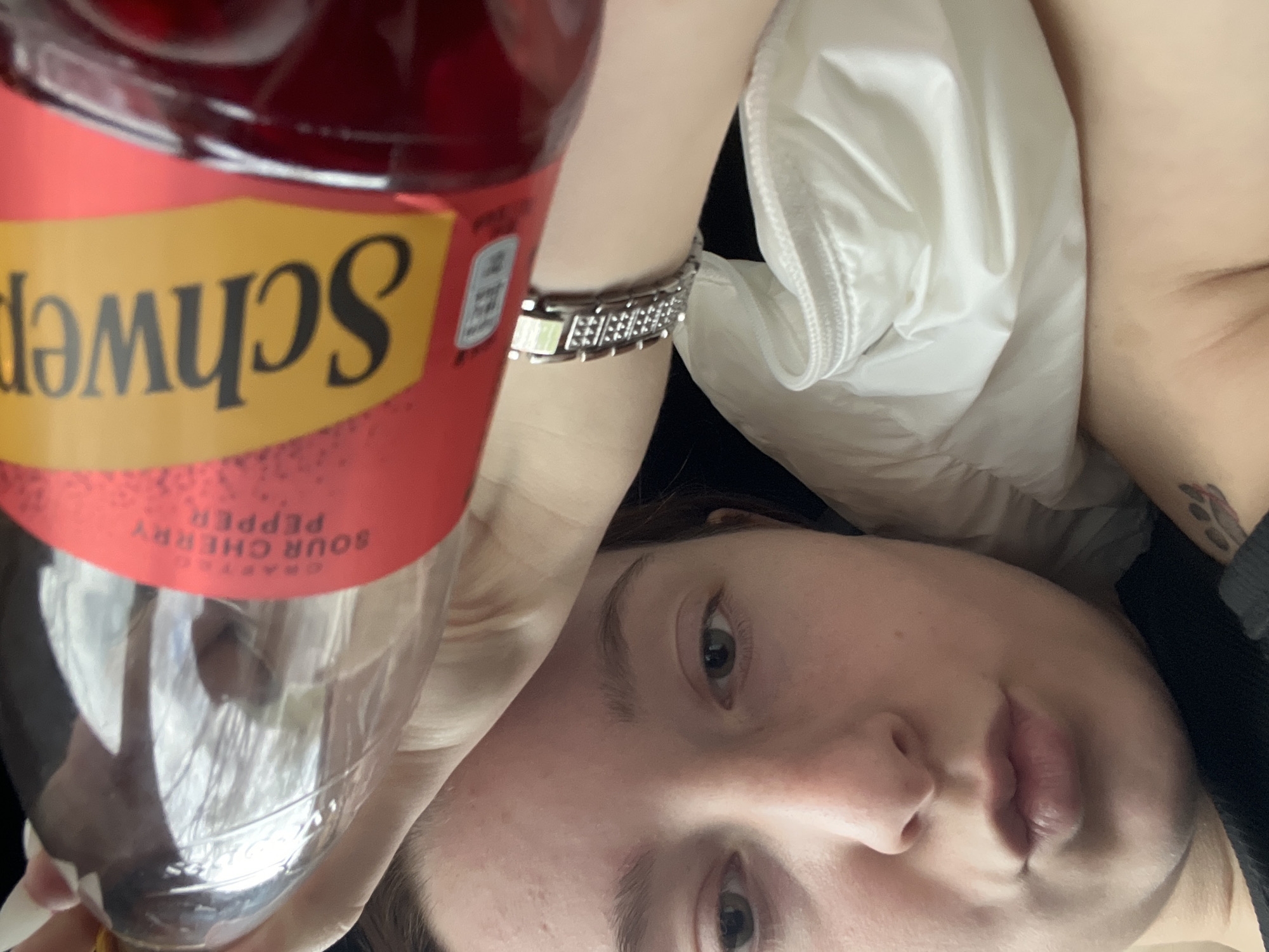 The image shows a person holding a bottle of Schweppes Crafted Sour Cherry Pepper in their left hand. The person is lying down, possibly on a bed, with a white puffy material (likely a jacket or blanket) visible next to them. The person is wearing a silver bracelet on their left wrist. The person's face is visible, showing their eyes, nose, and lips. The person also has a tattoo on their upper arm. The background is dark and out of focus. There is no information in the image that indicates it was taken in Varna, Bulgaria.
