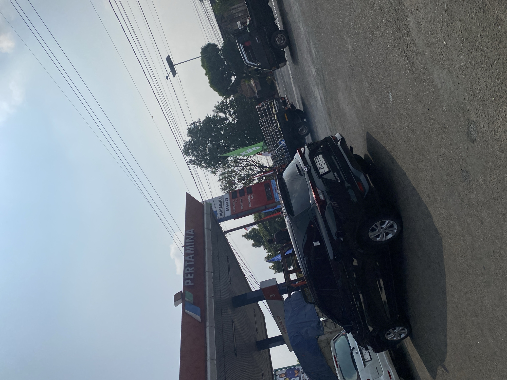 The image depicts a Pertamina gas station in Mantren, Indonesia, under a clear, bright sky with visible power lines. The gas station features a red awning with the Pertamina logo prominently displayed. A black SUV is parked in the foreground, facing away from the camera, with its license plate visible. To the left, a white vehicle is parked. Several other vehicles, including a black truck with a metal cargo bed, are on the road or at the gas station. Green and white flags are visible, as are signs indicating fuel types and prices. The environment appears to be a street scene with trees and buildings in the background.