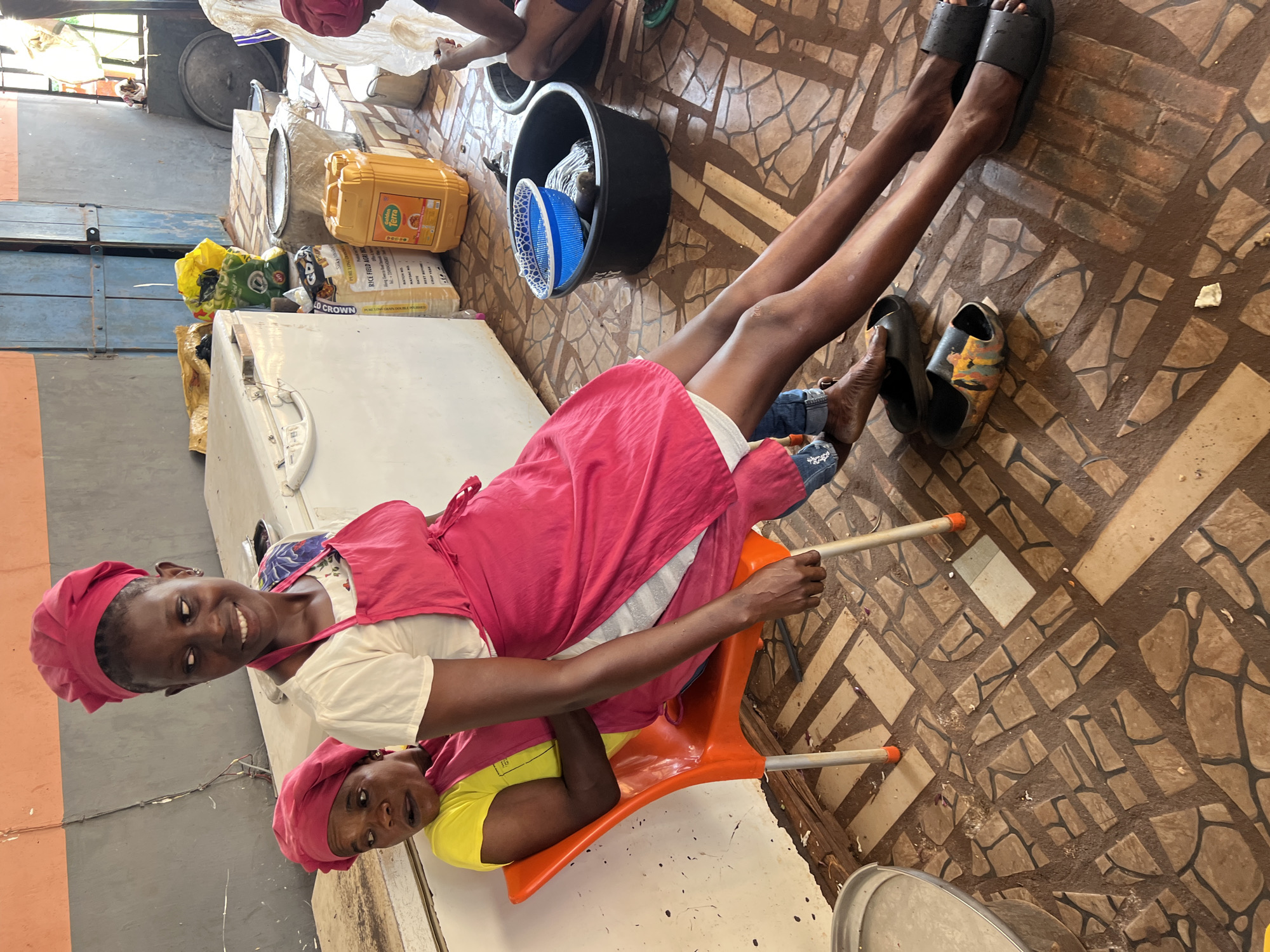 Captured indoors, the scene unfolds on a geometrically patterned tile floor, presumably within a food preparation area in Obosi, Nigeria. The space is occupied by two women, both wearing matching pink head coverings. One woman, donning a pink apron over a white top, sits on an orange chair, while the other, in a yellow shirt, leans against her. The sitting woman's legs extend, revealing black and patterned slides. Bags and containers of food products, including rice and cooking oil, are placed near a white refrigerator. Additionally, there's a black bucket and a blue colander nearby, adding to the context of food preparation.
