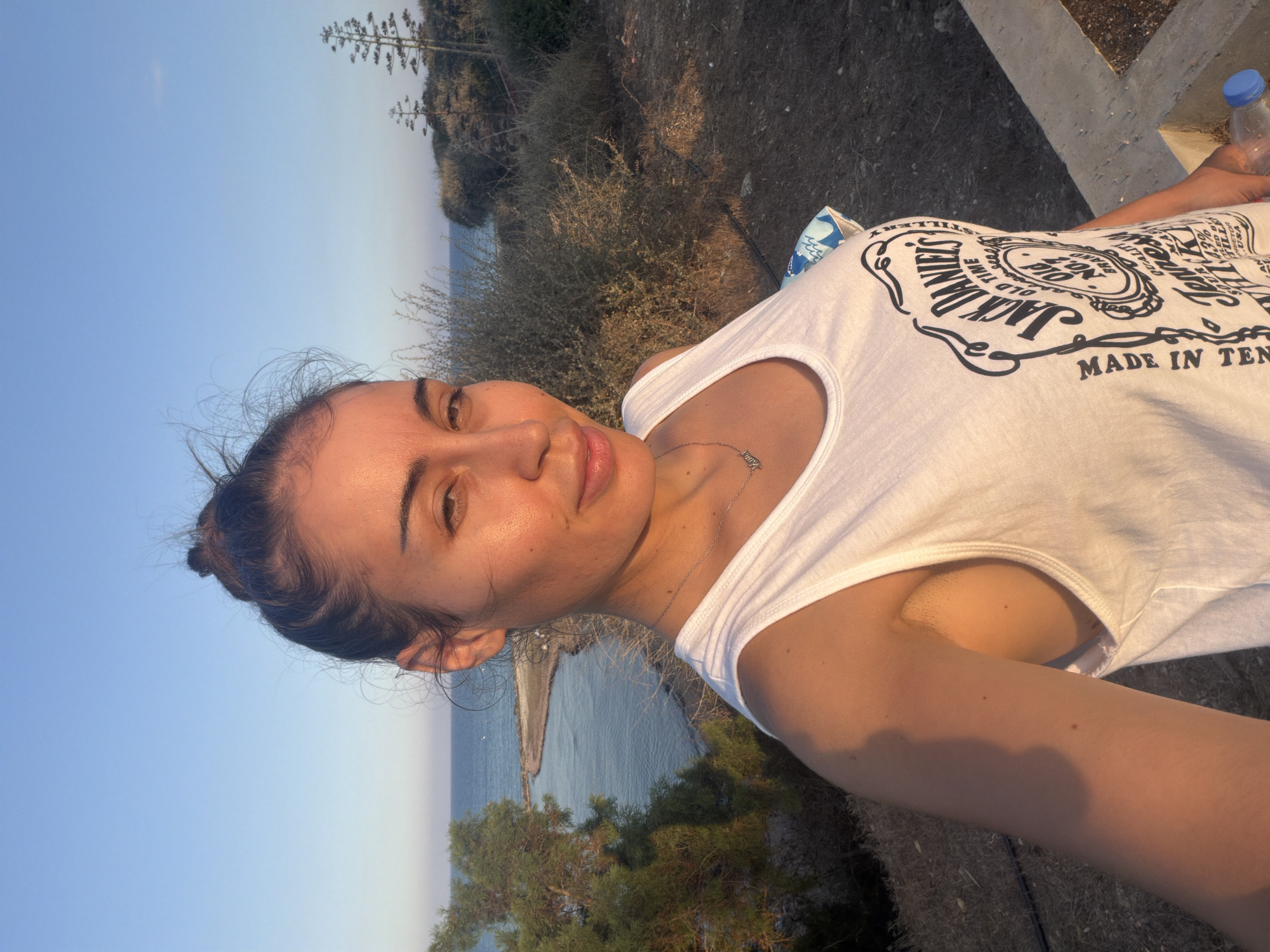 A woman with dark hair pulled up into a bun and light skin is taking a selfie outdoors with a body of water and vegetation in the background. She is wearing a white tank top with a Jack Daniel's Old No. 7 Brand Tennessee Whiskey logo printed on the front. A silver necklace with a nameplate is visible around her neck. The setting appears to be a coastal area with dry brush and a few trees, possibly in or near Meneou, Cyprus, given the clear blue sky and natural landscape. The sunlight suggests it is late afternoon or early evening.