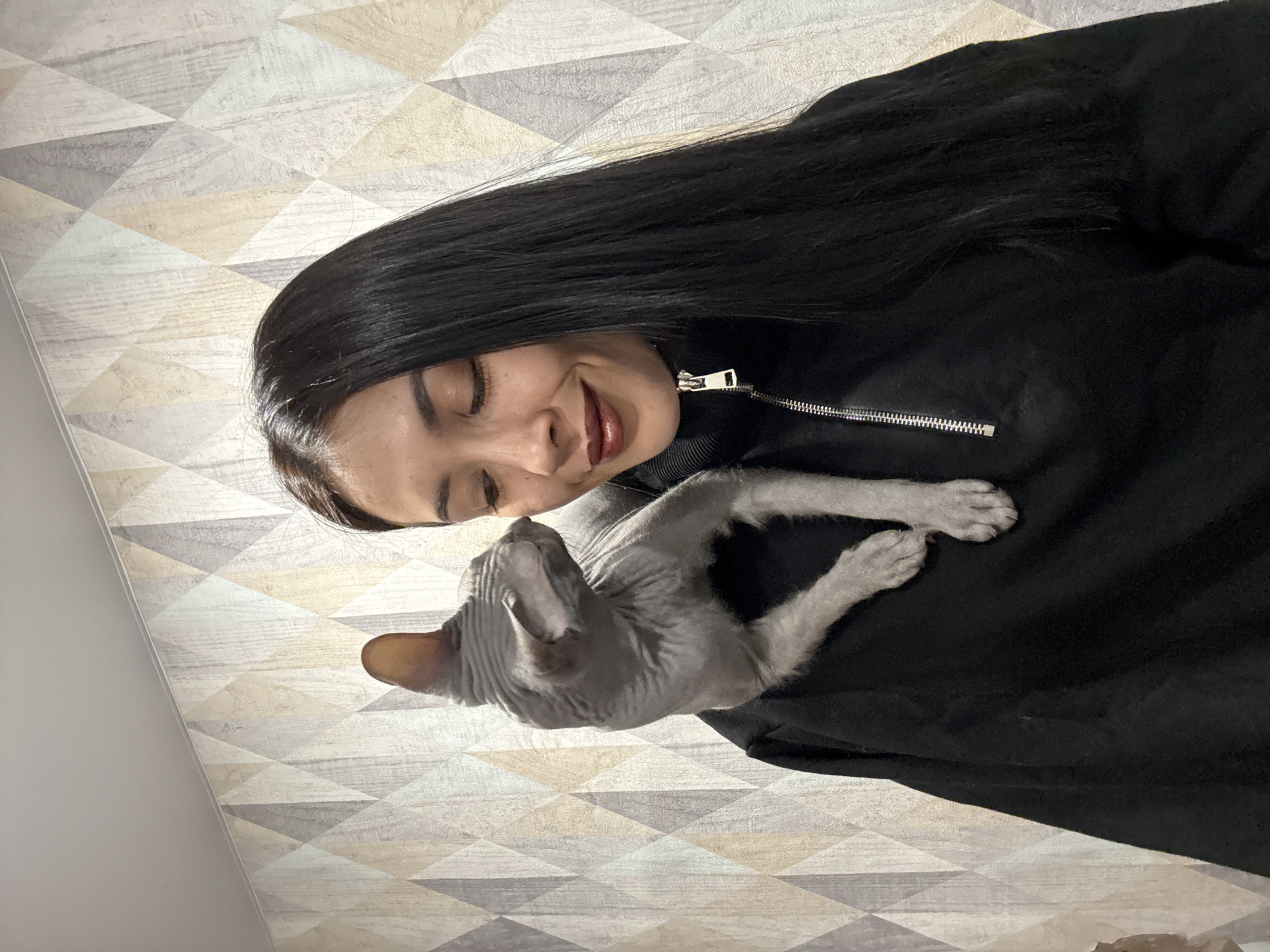 A woman with long, dark hair smiles gently down at a hairless grey cat resting on her chest. She wears a black top with a silver zipper detail near her shoulder. The background is a wallpaper with a geometric pattern consisting of triangles in shades of grey, white, and beige. The scene is indoors. There is no information provided that connects the image to the city of Votkinsk, Russia, so the location cannot be confirmed.