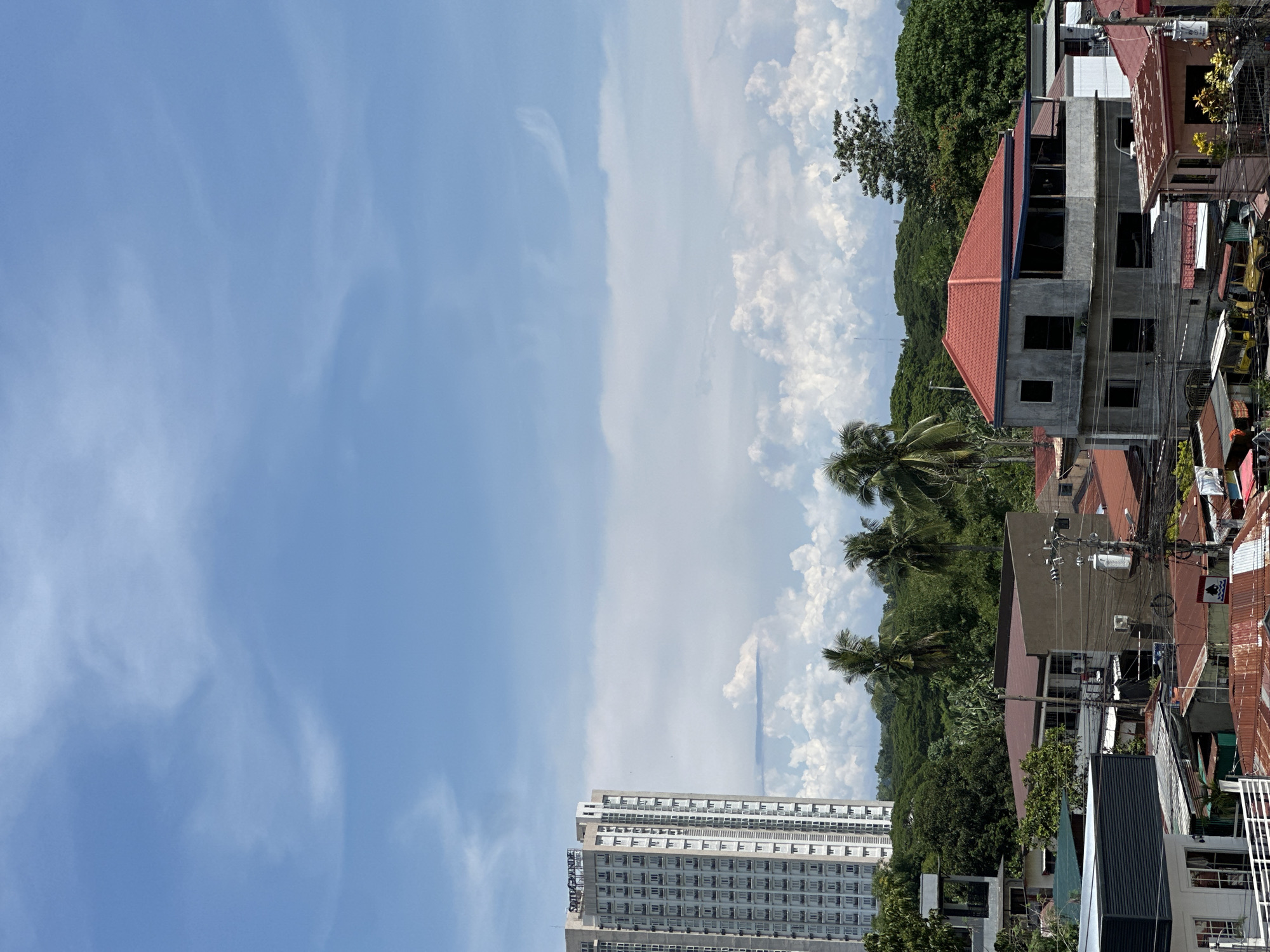 The image captures an urban scene in Davao City, Philippines. In the foreground, there's a partially-constructed building with a red roof amidst a dense cluster of older, residential structures with rusted roofs and an array of electrical wires. Palm trees and lush greenery intermingle with the urban landscape. In the distance, a tall, modern building rises prominently against a backdrop of a partly cloudy sky. The sky transitions from a clear blue on the left to a cloudier, whiter expanse toward the right.