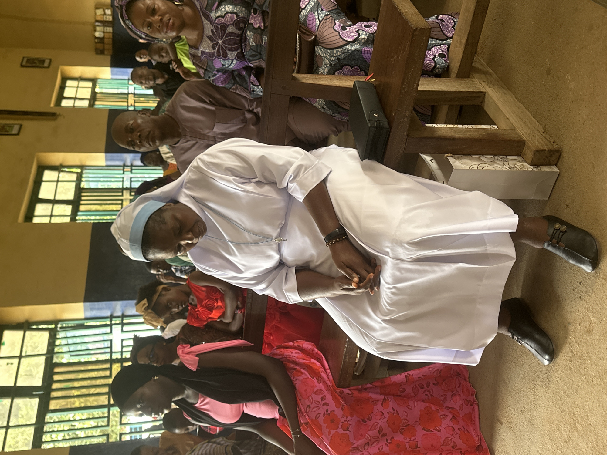 This media file shows a group of people, primarily women, seated in what appears to be a church or community hall in Akwanga, Nigeria. The image is oriented vertically, suggesting it was taken with a phone.
In the foreground, a woman in a white religious habit and head covering is seated. She is wearing dark shoes and has her hands clasped in her lap. Behind her, other individuals are visible, including a woman in a colorful patterned fabric and a man in a light brown traditional outfit. The setting includes wooden benches and windows with colorful panes, characteristic of a religious or communal gathering space. There are framed pictures on the wall and what looks like a Bible or religious book on a nearby wooden structure.
The people are attentively seated, suggesting a service, sermon, or meeting is in progress. The overall mood seems calm and respectful. The lighting suggests it is daytime, and the indoor setting implies no specific weather conditions are directly relevant. The presence of traditional Nigerian attire and the given location context of Akwanga, Nigeria, provide strong clues about the cultural setting. No visible text is discernible in the image.
