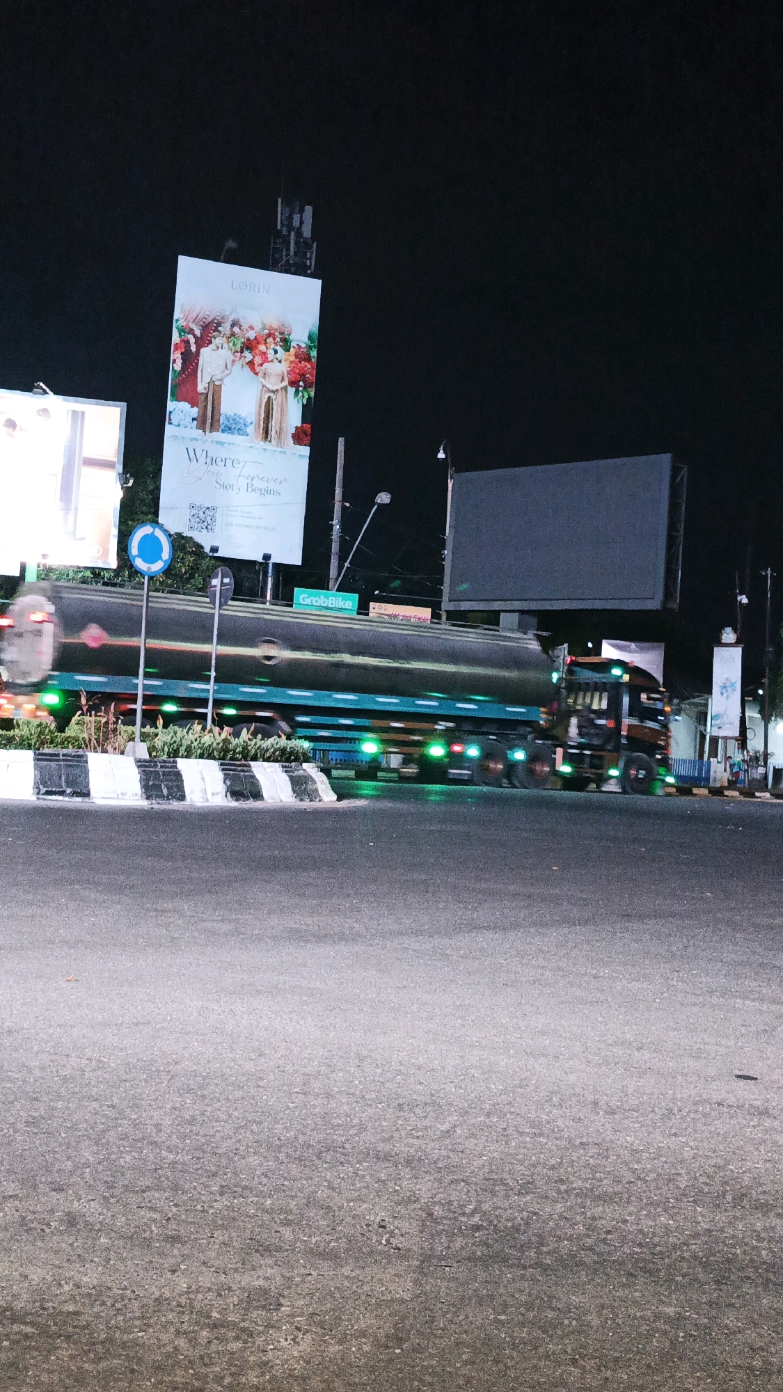 A long shot captures a tanker truck in motion at night on a street in Surakarta, Indonesia, suggesting a commercial area given the presence of multiple billboards. The truck's metallic tank reflects the neon green lights from its underside and the surrounding environment. In the background, a large billboard advertises "LORIN" with an image of a couple in formal attire and floral decorations, accompanied by the text "Where Your Forever Story Begins." Another large, dark billboard is visible to the right. Smaller signs, including one for "GrabBike," are also present. The road is asphalt, and a median strip with white and black striped barriers is situated in the foreground. The scene is illuminated by artificial lights from the billboards and the truck, with the sky appearing dark, indicating nighttime.