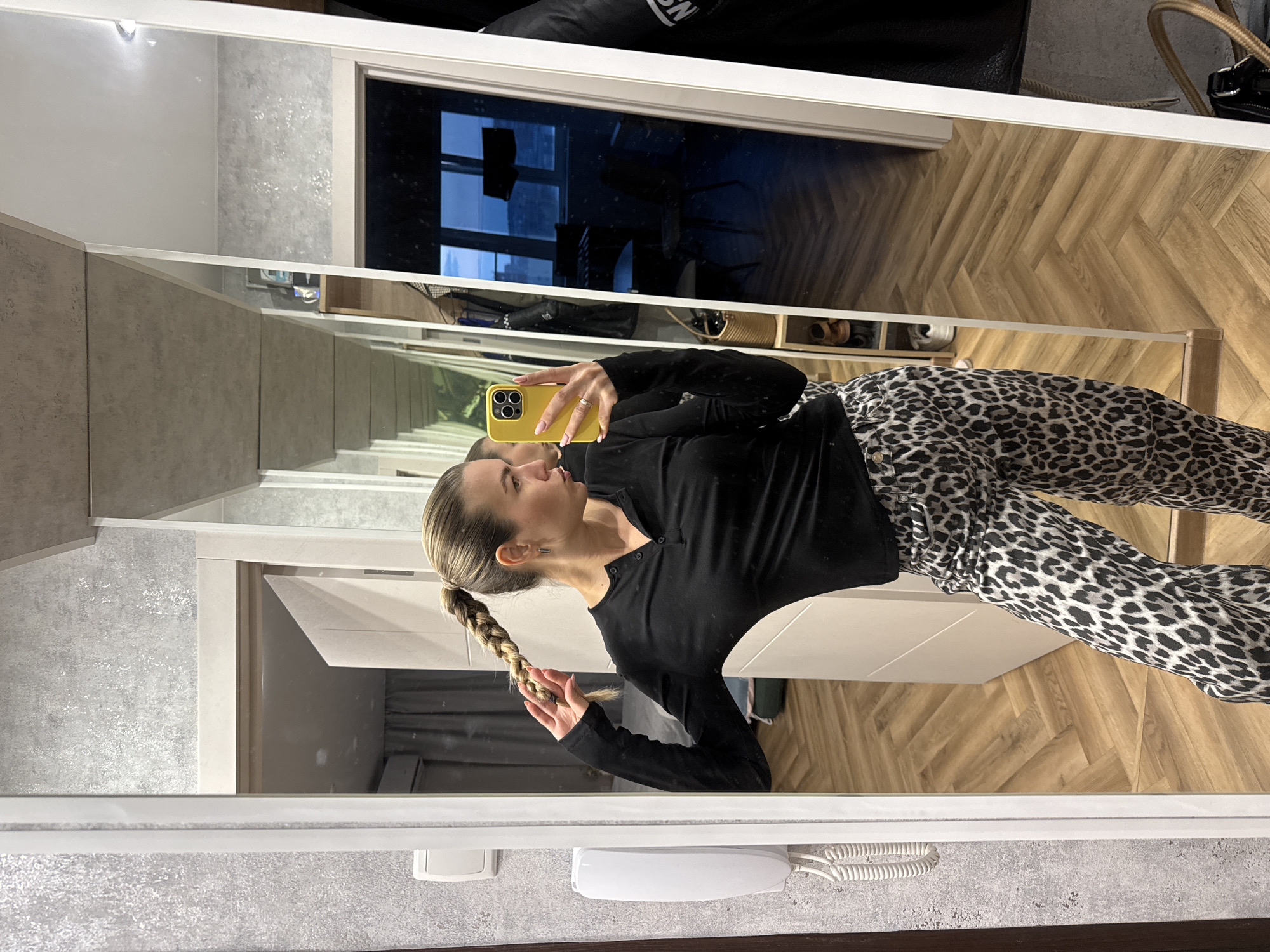 A blonde woman with her hair in a braid poses for a mirror selfie in a room with a herringbone wooden floor. She is wearing a black long-sleeved shirt and leopard print pants. She is holding a yellow smartphone in her right hand, which is angled to capture her reflection. The room appears to be a bedroom or dressing area, with a wardrobe, a window with a city view in the background, and a wall-mounted phone on the left side of the frame.