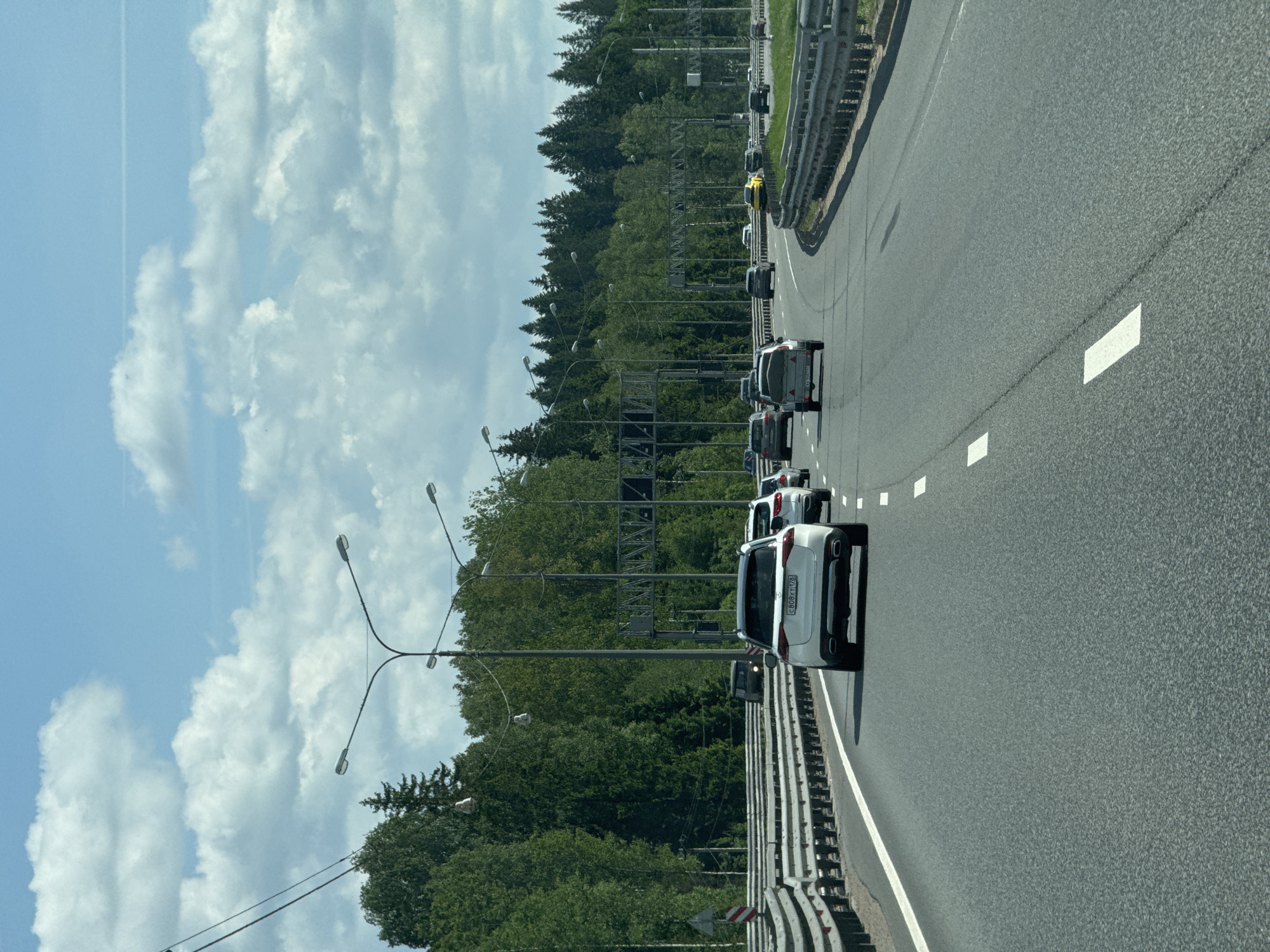 A daytime view captures a multi-lane highway near Vartemyagi, Russia, under a bright sky with scattered white cumulus clouds. The perspective is from within a vehicle, looking forward down the road. A line of cars is visible, indicating moderate traffic or a busy road.
In the immediate foreground, the rear of a white crossover or SUV with a license plate reading "c808xt178" is prominent. Ahead of it, several other vehicles, including a dark-colored sedan and other SUVs, are queued. Further down the road on the right, a distinctive yellow sports car is partially visible. The asphalt road surface is clearly detailed with white lane markings.
To the left of the highway, a dense forest of green trees lines the road, creating a natural boundary. Interspersed within the trees and along the roadside are tall streetlights with multiple arms, illuminating the area. A metal guardrail runs along the right side of the road, separating it from the adjacent terrain, and another appears on the left further back. A faint white contrail from an aircraft is visible in the distant blue sky. An overhead gantry structure for signs or cameras can be seen further down the road above the lanes. A small, red and white chevron road sign is partially visible near the guardrail on the left.