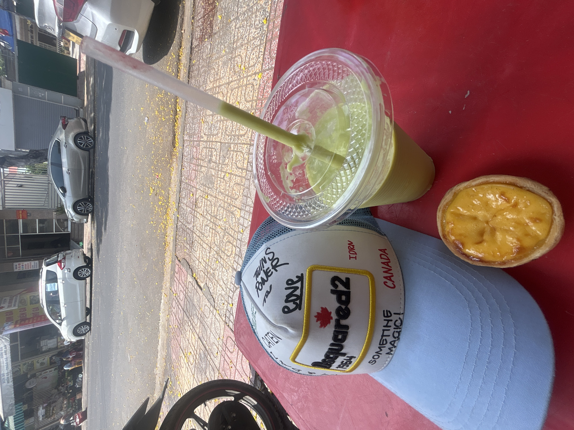 On a red surface, likely an outdoor table in Bac Natural Heritage Area Trang, Vietnam, a plastic cup with a green smoothie and a straw rests next to a small tart. The cup has a clear plastic lid, and the smoothie appears to be made with tropical fruits. Next to the tart sits a light blue baseball cap with a white front panel, featuring the brand name "Dsquared2" prominently displayed along with other text and a maple leaf emblem. The cap also has "Canada" and "SOMETIHE MAGIC!" printed on it. Part of a motorcycle tire is visible in the bottom left corner, suggesting the table is roadside. A street scene is blurred in the background, showcasing vehicles and buildings, indicating the location is an urban setting.
