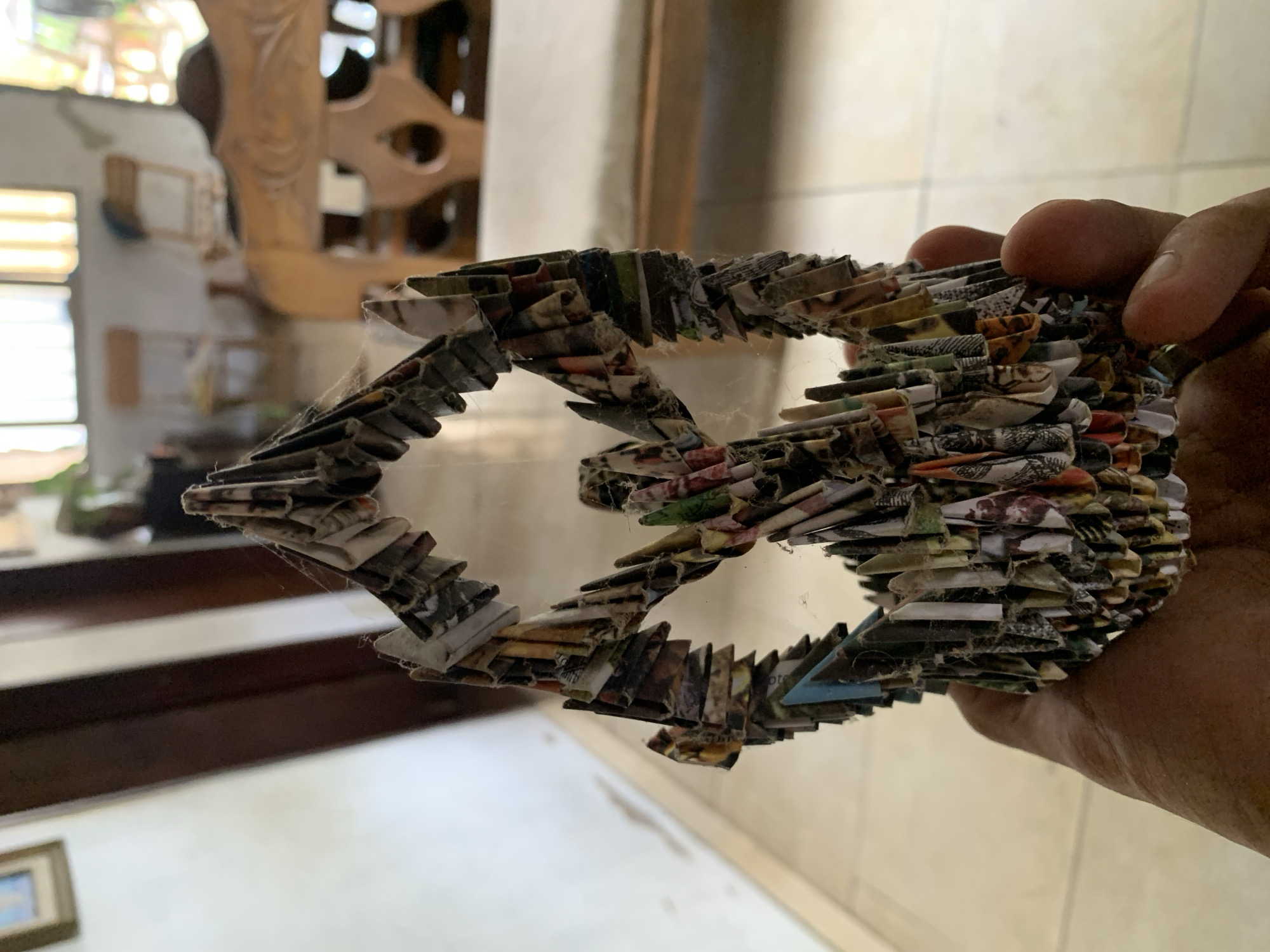 A person is holding a homemade fish-shaped ornament made from layered, rolled paper. The ornament has some cobwebs visible within its structure. The person's hand holding the ornament is tanned. The background shows a staircase, some wooden furniture, and a window, suggesting an indoor setting. The exact location in the city of Santa Ana, Venezuela is indeterminable without further information or context.
