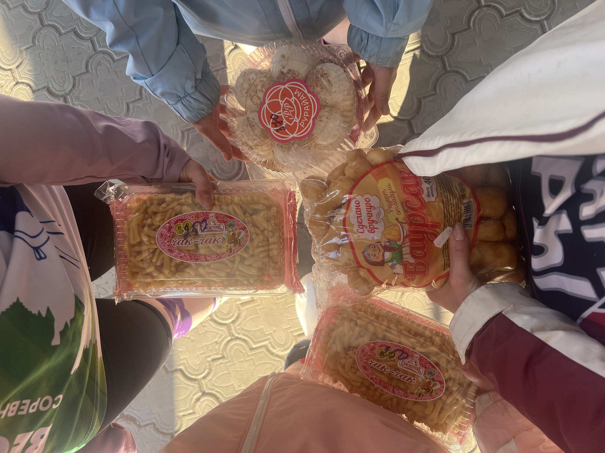 Four individuals are holding various packages of what appear to be traditional Tatar or Bashkir sweets. The sweets include "chak-chak" and "baursak". The location may be near Nizhnie Yaki, Russia, as indicated on the packaging. The individuals are wearing casual clothing. The image is taken from a top-down perspective, focusing on the sweets being held. The environment is outdoors on a patterned surface.