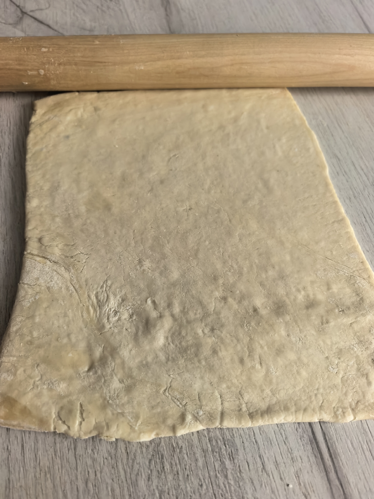 This close-up image captures a culinary scene, likely in a kitchen or baking area in Sterlitamak, Russia. The primary subjects are a light-colored, irregularly shaped piece of dough and a wooden rolling pin.
The dough, appearing to be pastry or bread dough, is spread flat on a light-colored surface, possibly a wooden countertop or table featuring a subtle gray and white wood grain pattern. It is an off-white or very light beige color, with a somewhat rough, uneven texture and slightly jagged edges. Traces of flour are visible on its surface, indicating the active process of preparation.
A smooth, cylindrical wooden rolling pin rests horizontally across the upper portion of the dough. It also shows faint dustings of flour, consistent with its use in flattening the dough. The rolling pin's position suggests it is either in the middle of being used or has just finished rolling the dough.
No people or visible text are present in the image. The scene conveys a sense of focused, quiet food preparation.