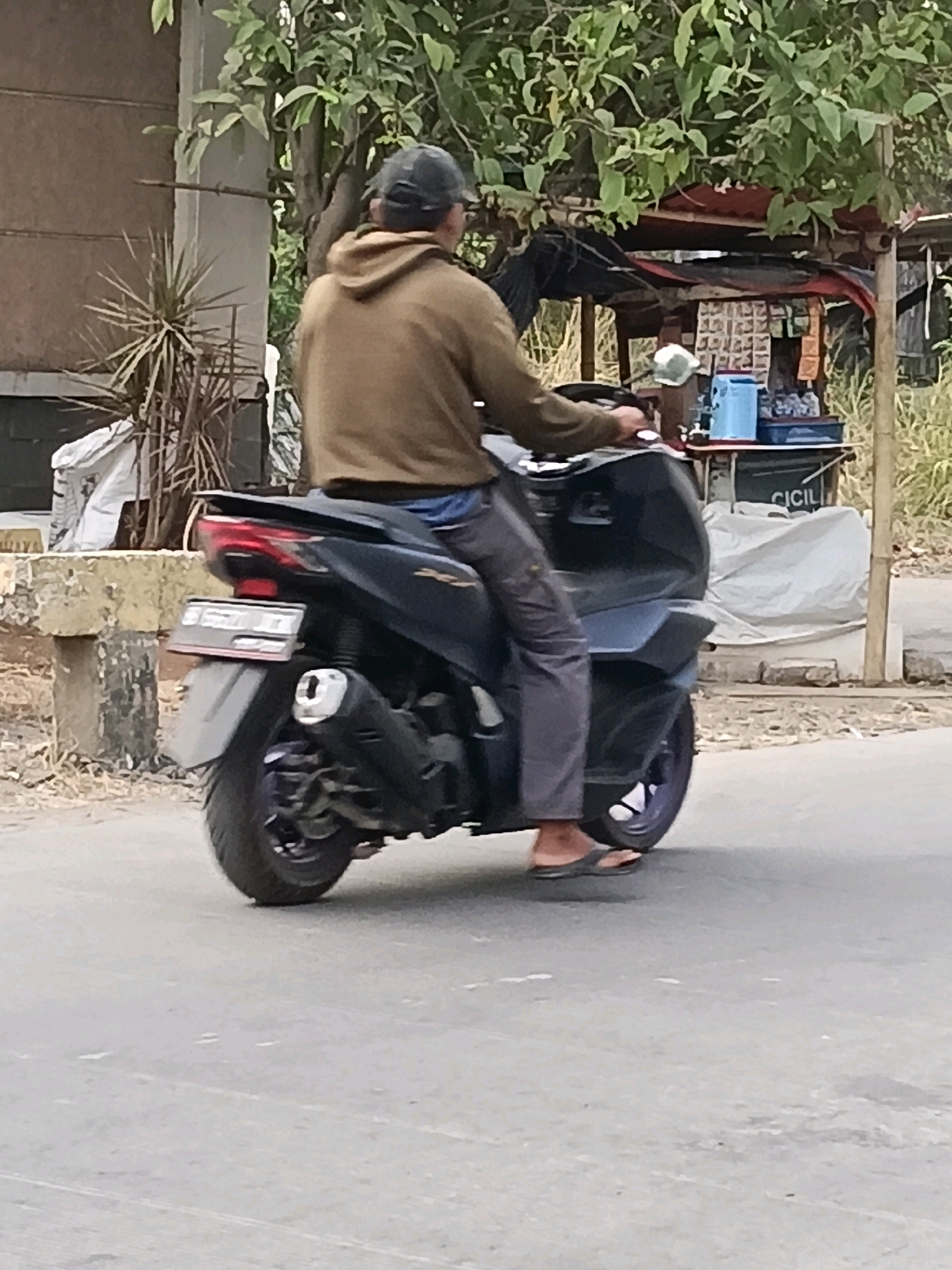 A person is riding a black scooter on a paved road in Belimbing, Indonesia. The person is wearing a brown hooded sweatshirt, gray pants, and flip-flops. They are also wearing a dark baseball cap. The scooter has a license plate that is partially visible. In the background, there is a building, trees, and a small roadside stall. The stall has various items displayed on shelves and a blue cooler. A large white tarp is draped over something behind the stall. The environment appears to be dry and dusty, with some dry vegetation visible.