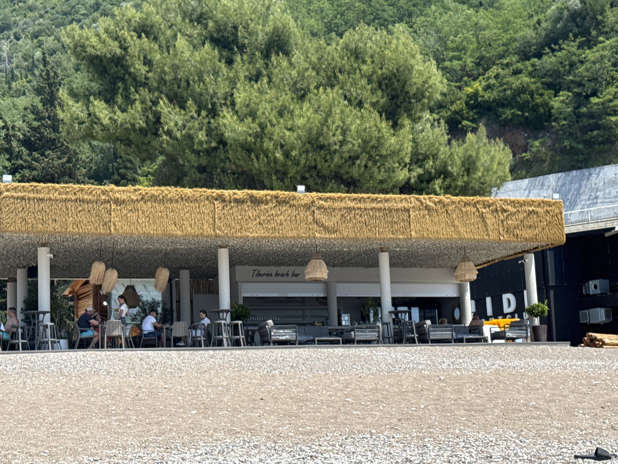 The image shows the Tiberina Beach Bar on a beach in Budva, Montenegro. The bar has a straw-covered roof supported by white pillars. Customers are seated at tables under the roof. Pendant lights hang from the ceiling. Behind the bar is a hillside covered with green trees. In front of the bar is a pebble beach. The lettering "IDI" is partially visible on the building to the right of the bar. People are sitting at tables, interacting with one another. The weather appears sunny.