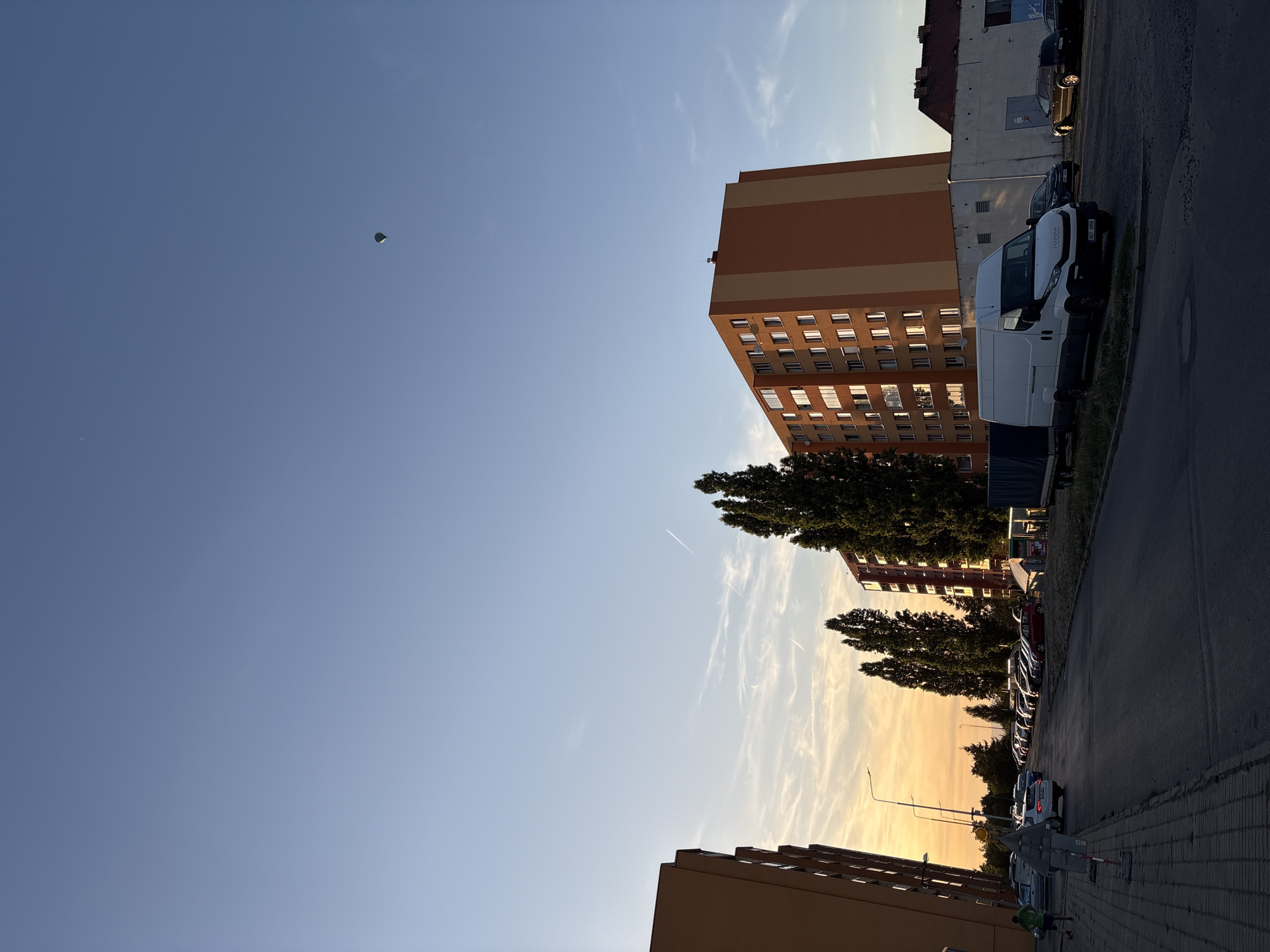 This is a medium-shot, eye-level, vertical, outdoor, full shot taken during the golden hour. The scene depicts a residential area with buildings, a street, parked cars, and a hot air balloon in the sky.
The sky is clear blue, with some wispy white clouds and faint contrails from airplanes. A small, dark hot air balloon is visible in the upper right quadrant of the sky. The sun is low on the horizon, casting a warm, orange glow on the clouds and the sides of the buildings.
In the foreground, there is a paved road with some parked cars. A white van with a dark trim and license plate that reads "907 995-B" is parked on the side of the road. To the left of the road, there is a sidewalk with a person wearing a green shirt and dark shorts running away from the camera. Further down the sidewalk, there are several parked cars.
Dominating the right side of the image is a tall, orange and beige apartment building with many windows. To the left of this building, there are several tall, dark green trees. On the far left of the image, part of another tall, orange apartment building is visible. Behind the apartment buildings, there are other structures, including a white building with a dark roof.
The overall impression is one of a quiet urban evening, with a hint of leisure activity suggested by the running person and the distant hot air balloon.
