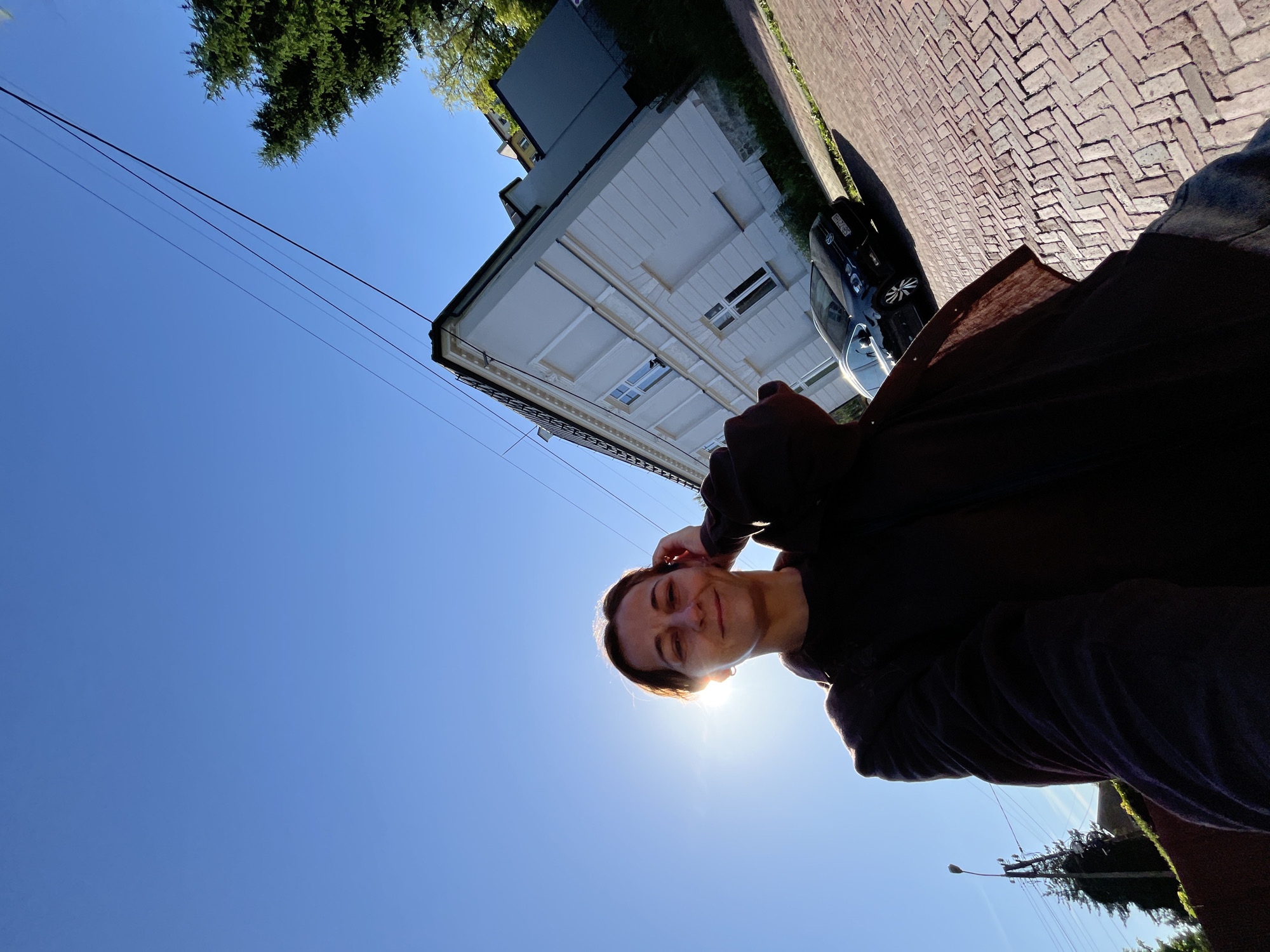 A low-angle, full-body selfie captures a woman standing on a brick-paved street in front of a light-colored building. The sun, positioned to the left of the frame, is partially obscured by the woman's head, creating a lens flare and backlighting her face. She is wearing a dark-colored jacket over a darker garment and denim jeans. Her right hand is raised, touching her hair near her temple. To her right, a dark-colored car is parked on the side of the street. The building behind her is two stories tall with white and light gray siding, featuring several windows. Overhead, power lines stretch across the clear blue sky, and a tall evergreen tree is visible on the right side of the frame. In the background, a lamppost and more trees are present.