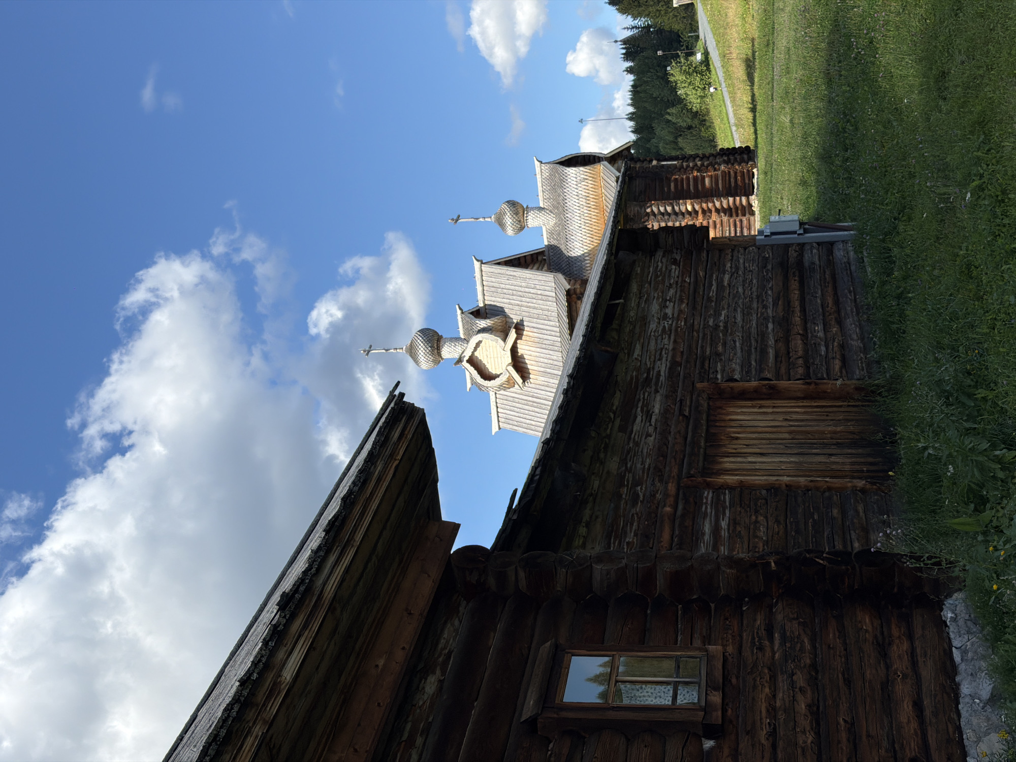The image depicts an outdoor scene in Gora (Khokhlovskoe S/P), Russia, under a bright blue sky with scattered white clouds. In the foreground, a weathered, dark wooden log building with a grey roof occupies the left side of the frame. A small window with a patterned curtain is visible on its facade. To the right of this building, two wooden structures resembling chapels or churches are visible, featuring distinctive wooden domes topped with crosses. The one in the foreground has a gray, textured roof, while the one behind it has a lighter, shingle-like roof. A pathway or road curves uphill to the right, bordered by green grass and trees in the background. A person is visible walking on the path in the distance. The overall environment suggests a rural or historical setting.