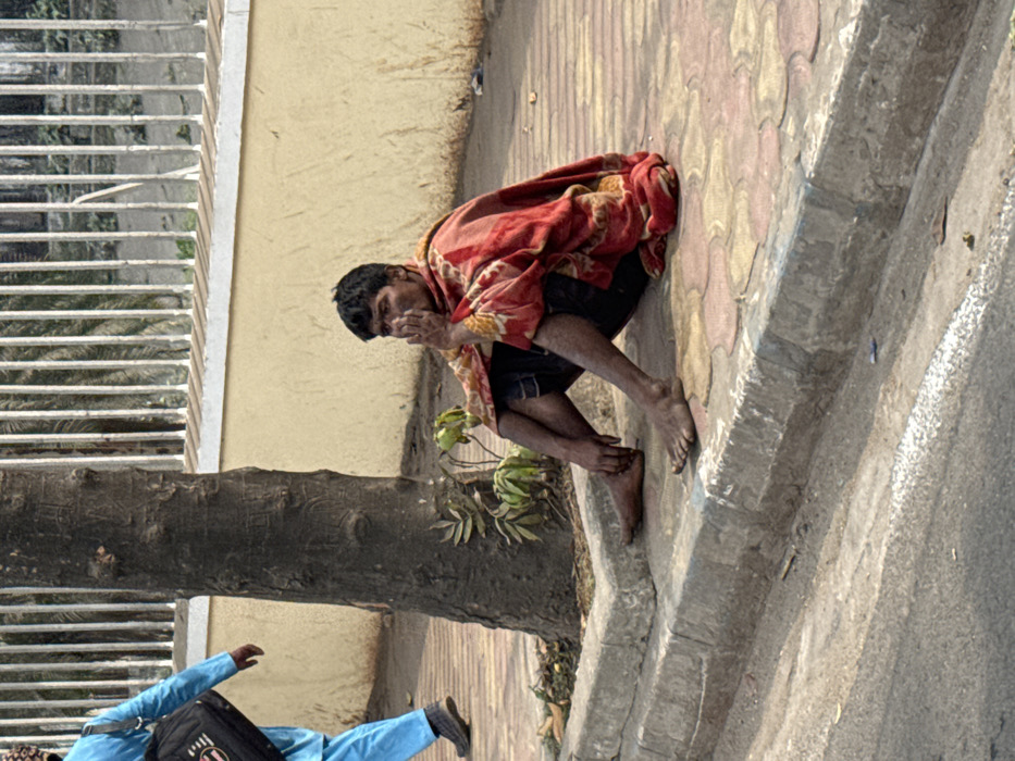 A person squats on a sidewalk in an urban setting, wrapped in a red and orange patterned blanket. Their dark hair contrasts with their skin tone, and they appear to be touching their face. Bare feet rest on the concrete edge of the sidewalk, adjacent to a tiled area. A tree trunk is visible to the left, alongside the lower portion of someone wearing blue clothing and carrying a black bag. A building with barred windows is in the background.