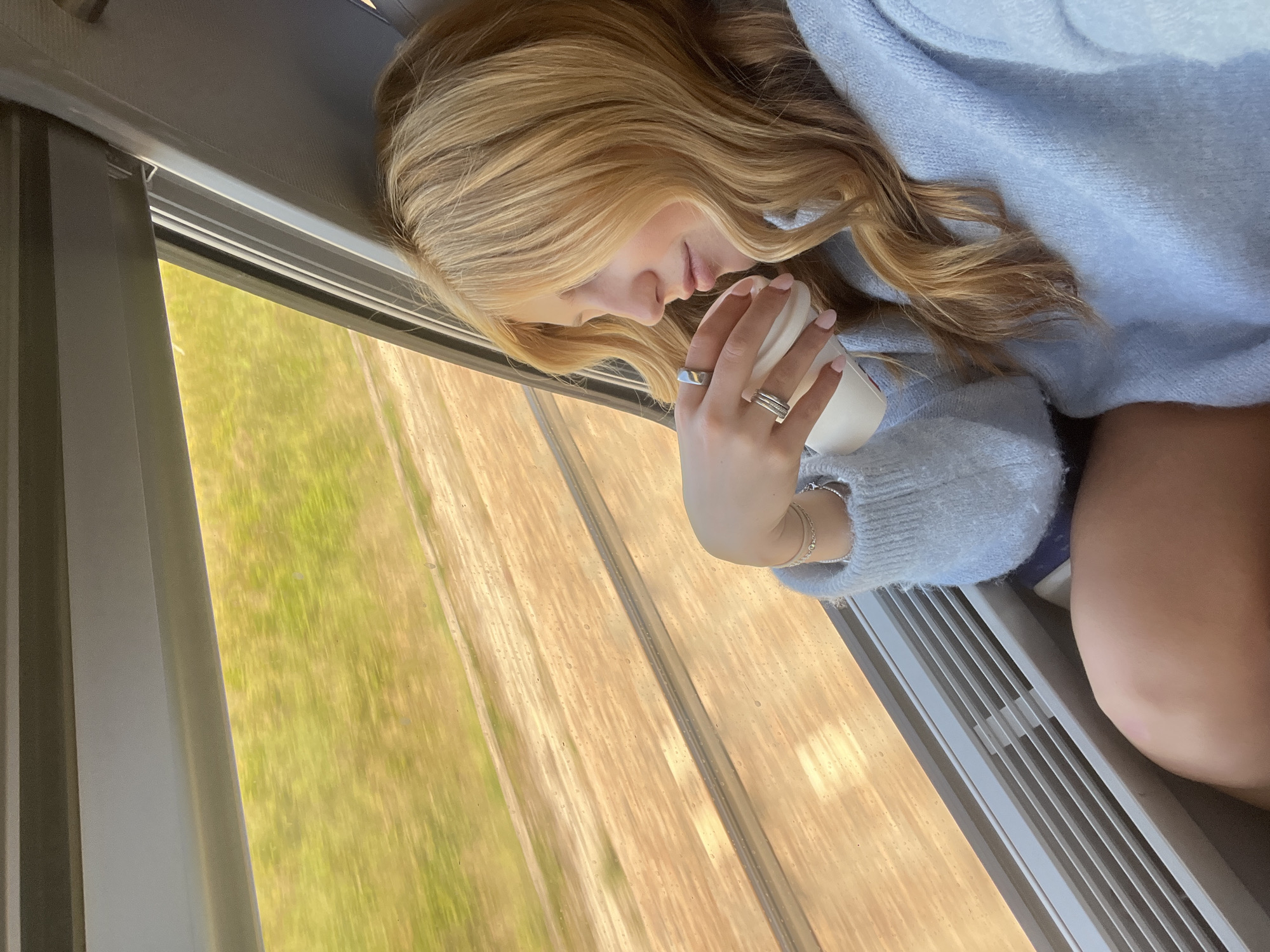 A young woman with long, wavy blonde hair is seen resting inside a moving vehicle, likely a train, given the context of Marmeaux, France. She is positioned horizontally, her head tilted down with eyes closed, suggesting she is sleeping or deeply relaxed. Her face is visible in profile.
She wears a soft, light blue knitted sweater. Her hand, adorned with multiple silver rings (one wide band and one with stacked parallel lines) and delicate silver chain bracelets, holds a plain white disposable cup. Her fingernails are manicured with light pink polish.
Through the window beside her, a blurred landscape of vibrant green fields and lighter brown earth or tracks rushes past, indicating rapid motion. The bright light from outside suggests it is daytime and sunny. The interior visible includes grey and white window frames and a section of horizontal slats, possibly an air vent, below the window. The overall atmosphere is peaceful and serene. No visible text is discernible in the image.