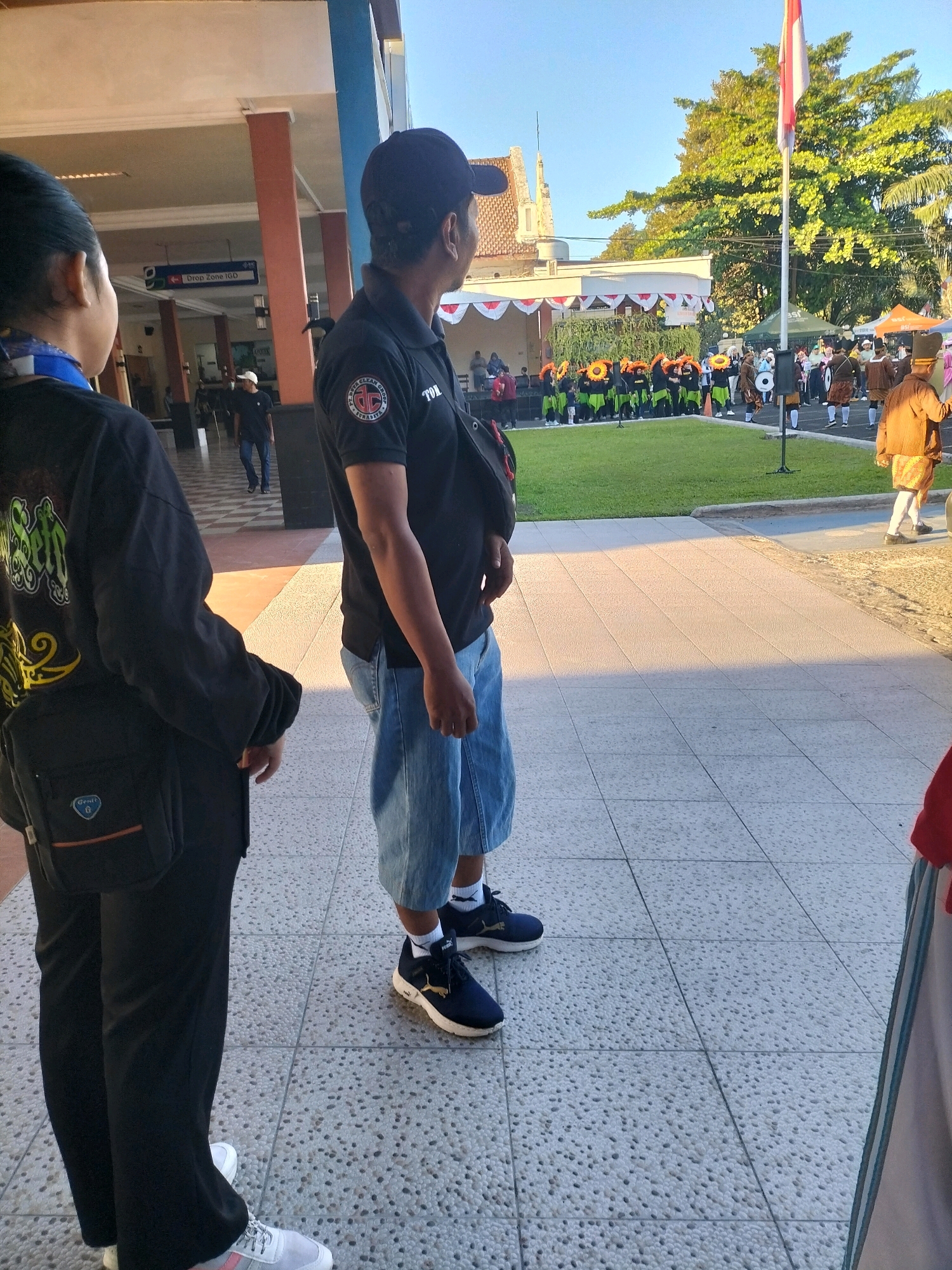 The image depicts an outdoor scene on a sunny day, likely in Pare, Indonesia, given the context. In the foreground, two individuals are visible from the waist down. The person on the left wears black trousers and white athletic shoes with pink accents, carrying a black messenger bag. The person on the right wears denim shorts and dark blue athletic shoes, with a black sling bag.
In the mid-ground, a group of people in uniform, possibly a marching band, are lined up on a grassy area. They are wearing green and orange costumes, and some are holding musical instruments. A flagpole with a red and white flag stands prominently behind them. Further in the background, there is a building with a covered walkway and architectural details suggesting a public institution, possibly a hospital or school, indicated by a sign that reads "Drop Zone IGD". The overall environment suggests a public event or ceremony taking place outdoors.