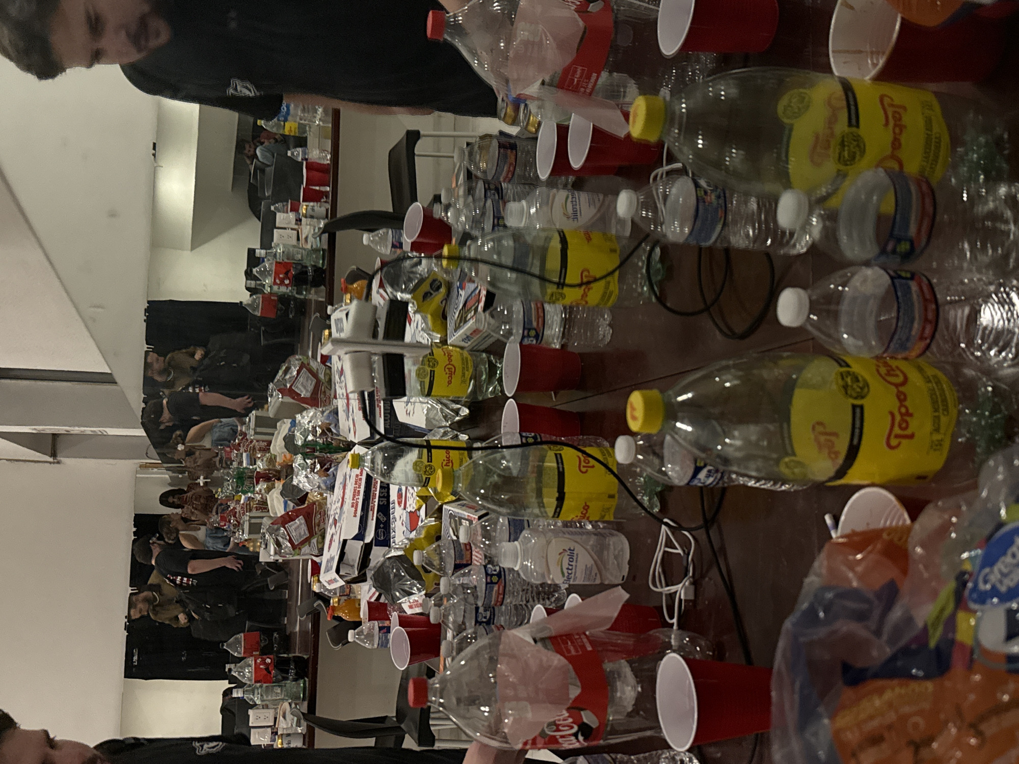 The image shows a table covered in empty bottles, red plastic cups, and food packaging. Empty Coca-Cola, Topo Chico, and Electrolit bottles are visible. Several people are reflected in a mirror behind the table, suggesting a gathering or party. The specific location in Tlalnepantla, Mexico is not discernible from the image.