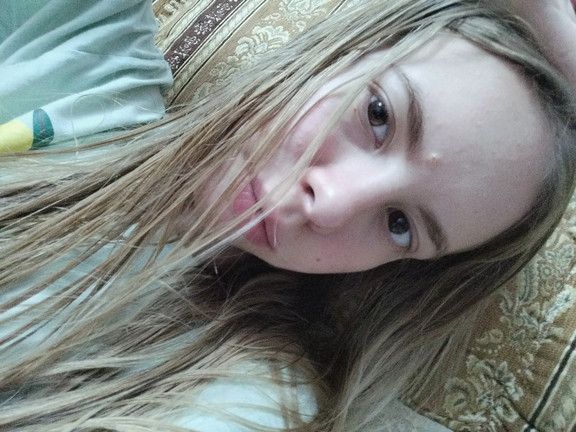 A close-up, low-angle, indoor selfie shows a young Caucasian female with long, blonde, slightly disheveled hair obscuring part of her face. She is lying down, with her head tilted, and her hair spreads across the frame. Her left eye is clearly visible, a dark brown iris, with subtle hints of pinkish blush on her cheek. Her lips are slightly parted, revealing a hint of her teeth. She appears to be wearing a light blue garment on her upper body, with a yellow and green striped detail visible on the left side of the frame. The background consists of a patterned fabric, possibly a sofa or bedspread, in shades of brown and cream with an ornate floral design. The lighting is soft and diffused, suggesting an indoor environment without direct sunlight. The overall impression is casual and intimate.