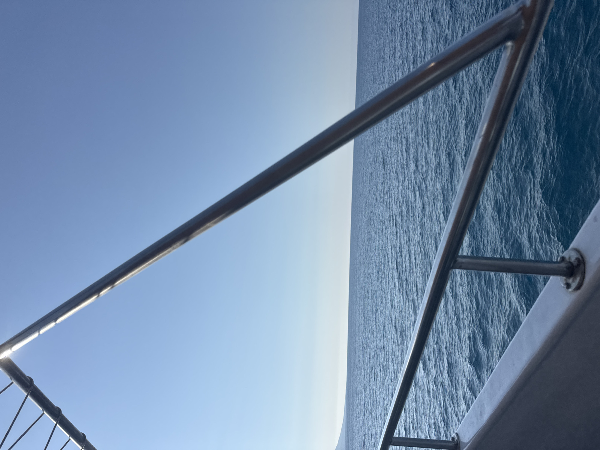 The image is a photograph taken from a boat on the sea. A metal railing is visible in the foreground, running horizontally across the bottom half of the frame and diagonally up the right side. The railing is attached to a light-colored surface, likely the deck of the boat. Beyond the railing, a vast expanse of deep blue ocean stretches to the horizon. The water is slightly choppy, with small waves visible. In the far distance, a faint landmass can be seen on the left side of the horizon, appearing as a low, dark line against the lighter sky. The sky is clear and a pale blue, gradually becoming lighter towards the horizon. The overall scene depicts a calm day at sea, with the viewer positioned on a vessel. The location is Neo Chorio, Cyprus, suggesting the boat is likely sailing in the waters off the coast of this Cypriot village, possibly near the Akamas Peninsula. There are no people or animals visible in the image.