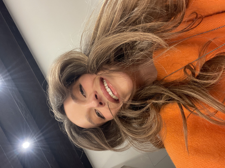 A woman smiles broadly in an upside-down selfie. Her long, wavy brown hair surrounds her face and spills across an orange sweater. A delicate chain necklace dangles amidst her hair. The background is a mix of light and shadow, with a bright spot illuminating a dark wall or surface to the left.